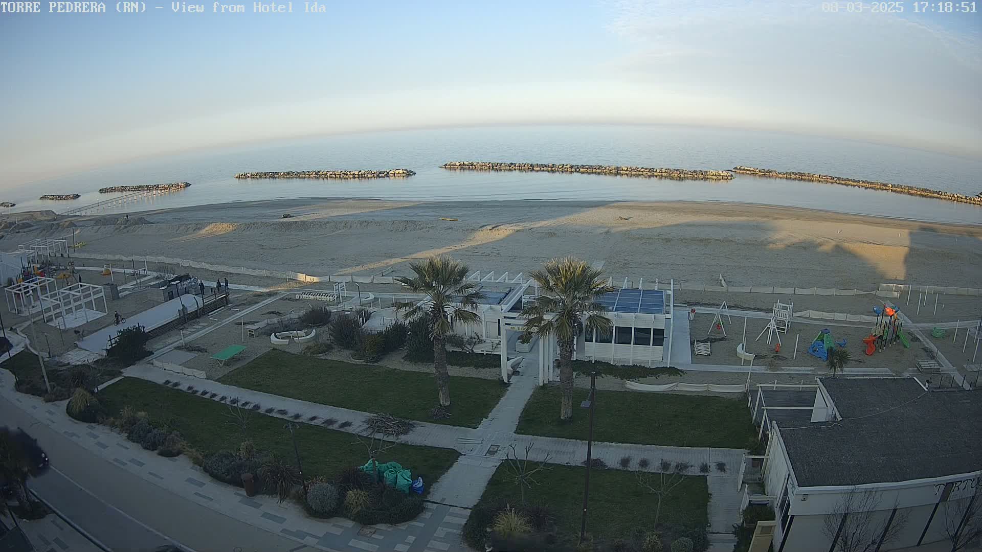 An aerial view shows a sandy beach with calm ocean water under a clear, bright sky; a playground, a small white building, and palm trees are visible near the beach.