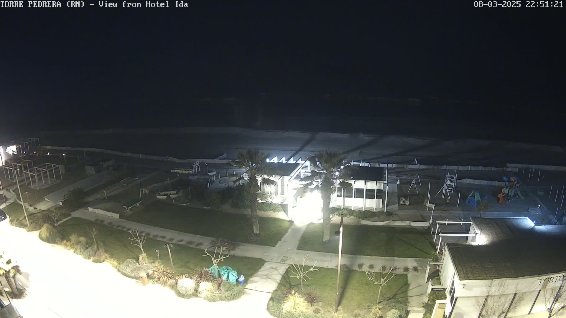 A nighttime, high-angle view of a beach area with a building, palm trees, a playground, and a mostly dark ocean.