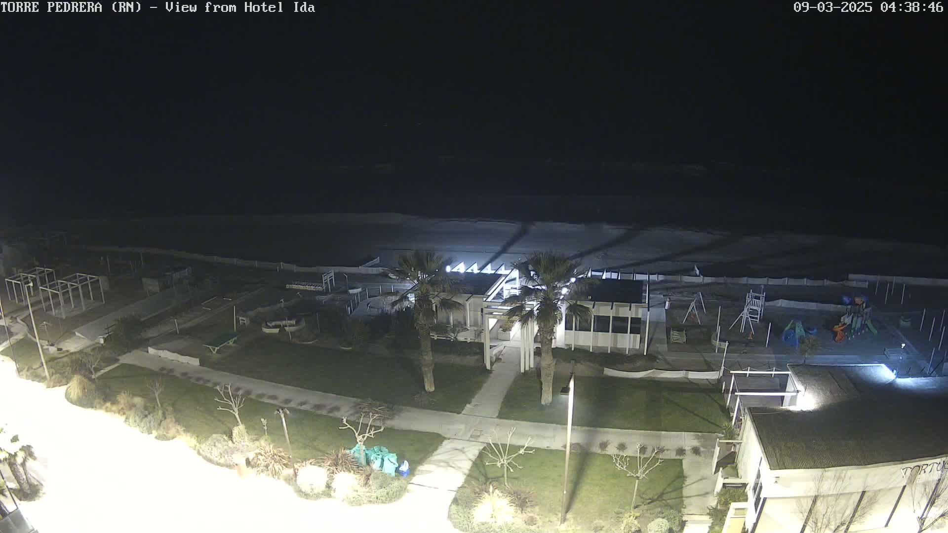A nighttime view of a beach, with a building, playground, and landscaping illuminated, overlooking a dark ocean under a clear sky.