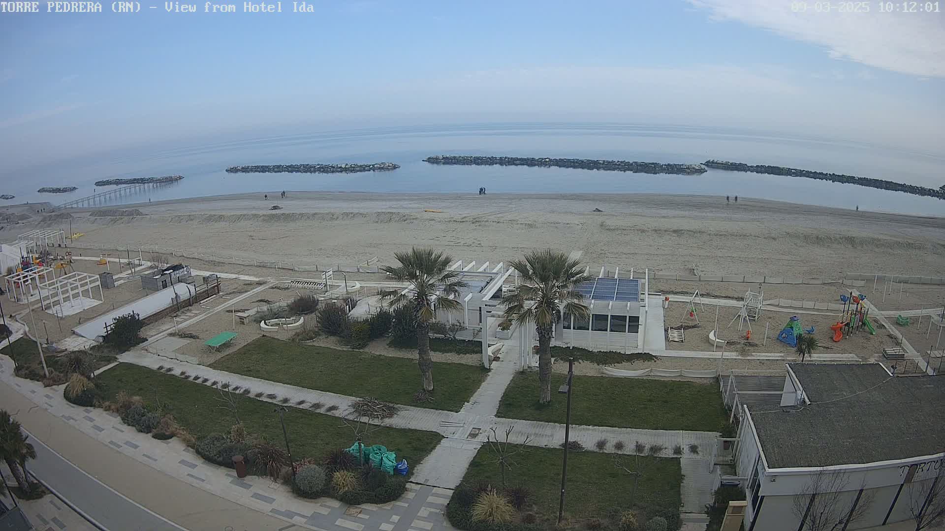 An aerial view of a beach with calm, overcast skies shows a sandy shore, a boardwalk, a playground, palm trees, and buildings.