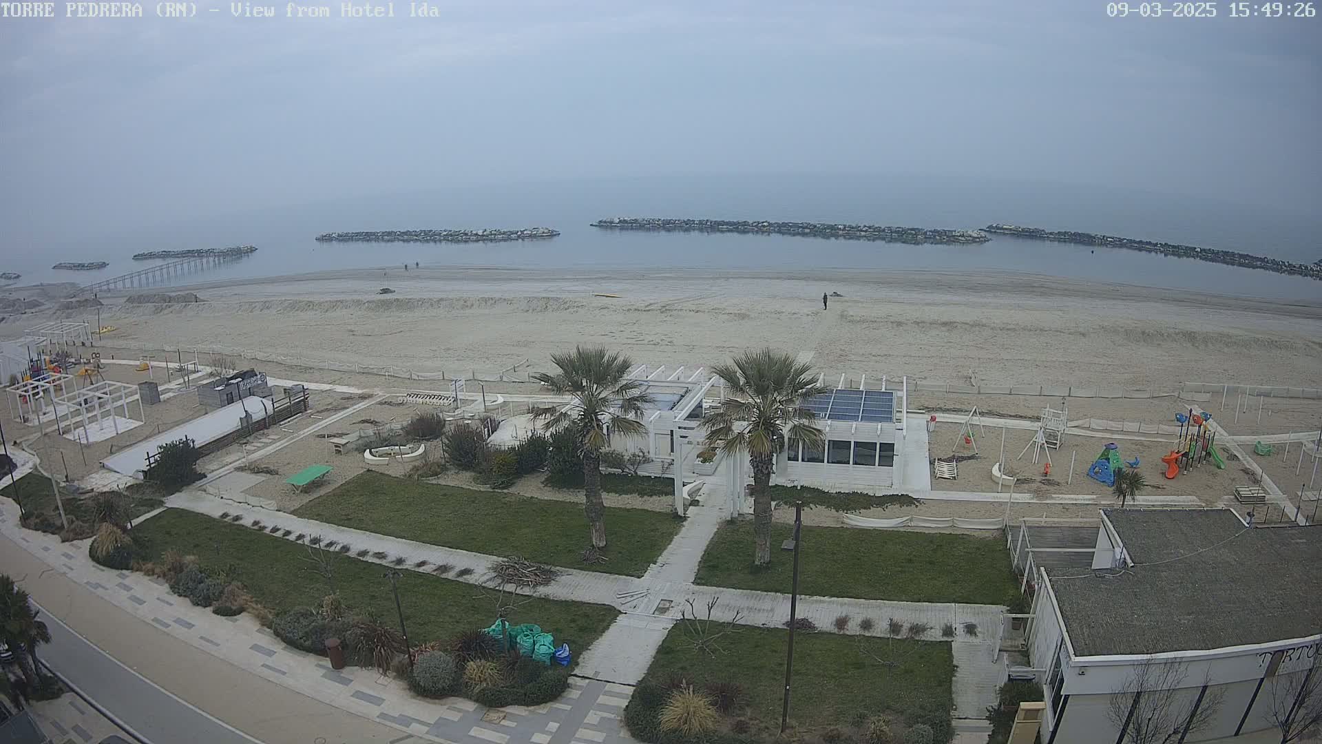 An overcast day reveals a sandy beach with a playground, a white building, and palm trees, all situated near a calm sea with breakwaters.
