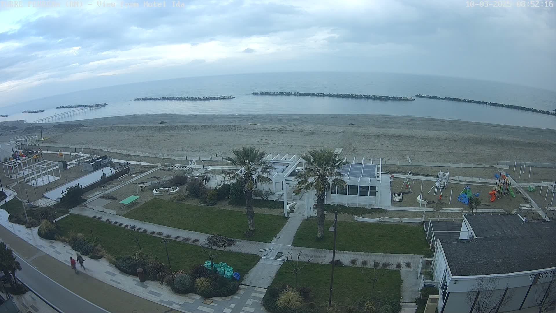 An overcast day reveals a beach scene with a sandy shore, calm sea, and beachfront structures including a playground, a white building, and palm trees.
