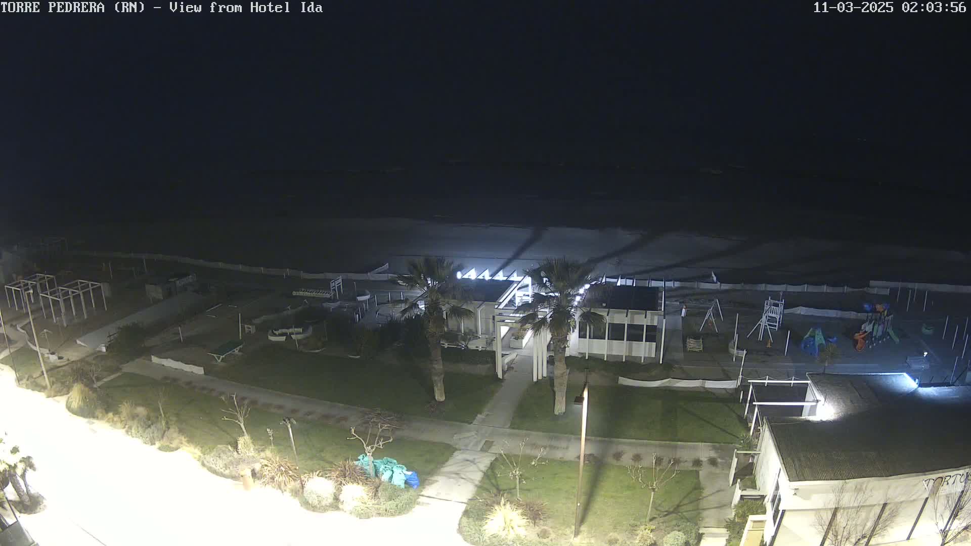 A nighttime, overhead view of a beach, playground, and beachfront building, illuminated by artificial lights under a dark sky.