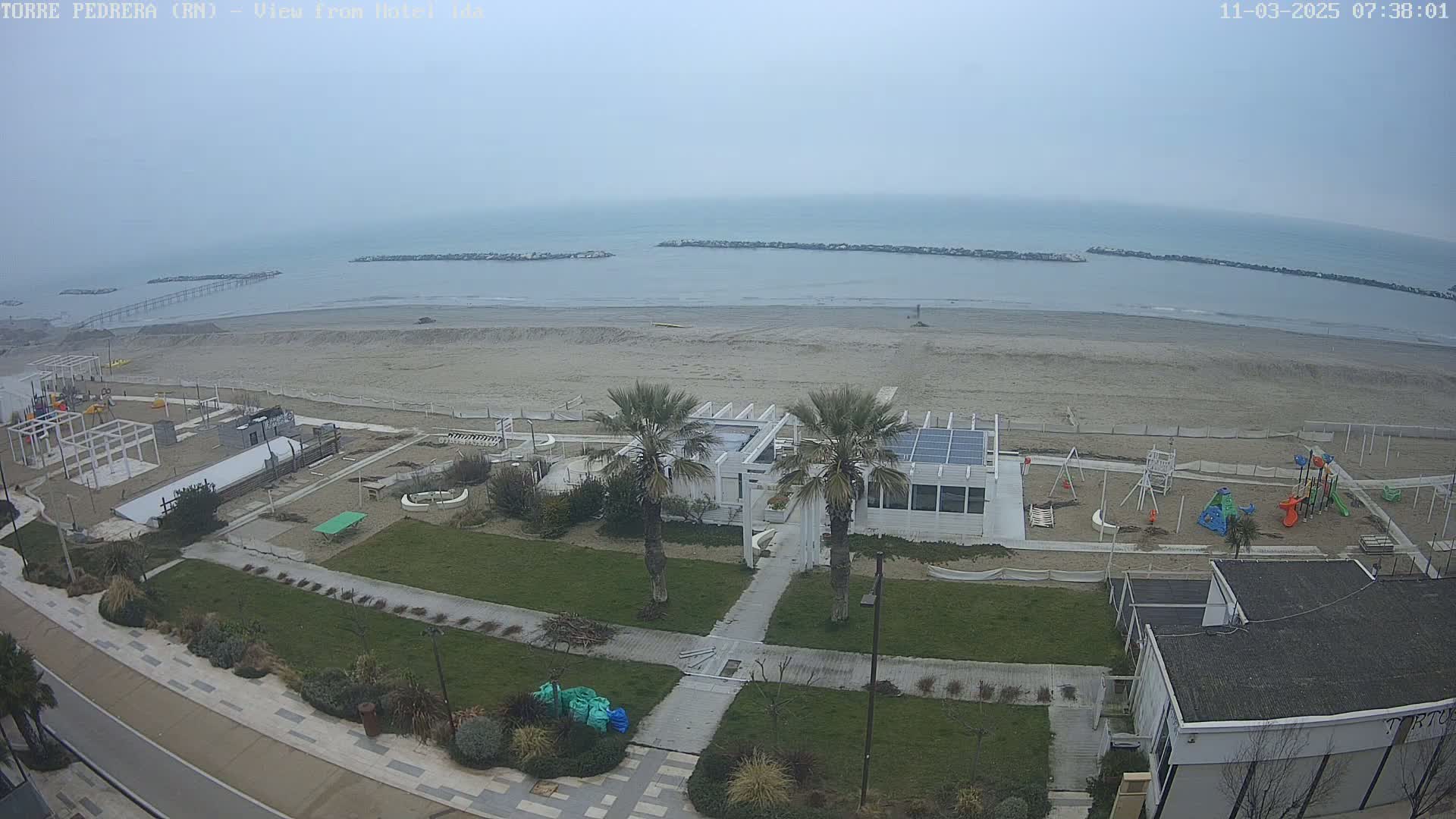 A beach with calm, overcast weather, featuring a playground, a beachfront building, and palm trees situated between the beach and a grassy area.