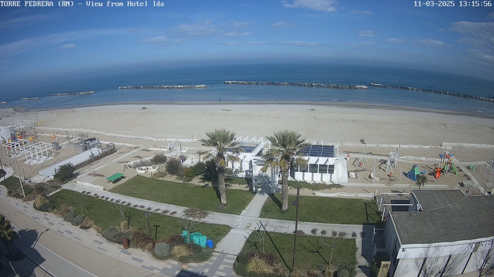 An aerial view of a beach with a playground, a building with solar panels, and palm trees on a sunny day.