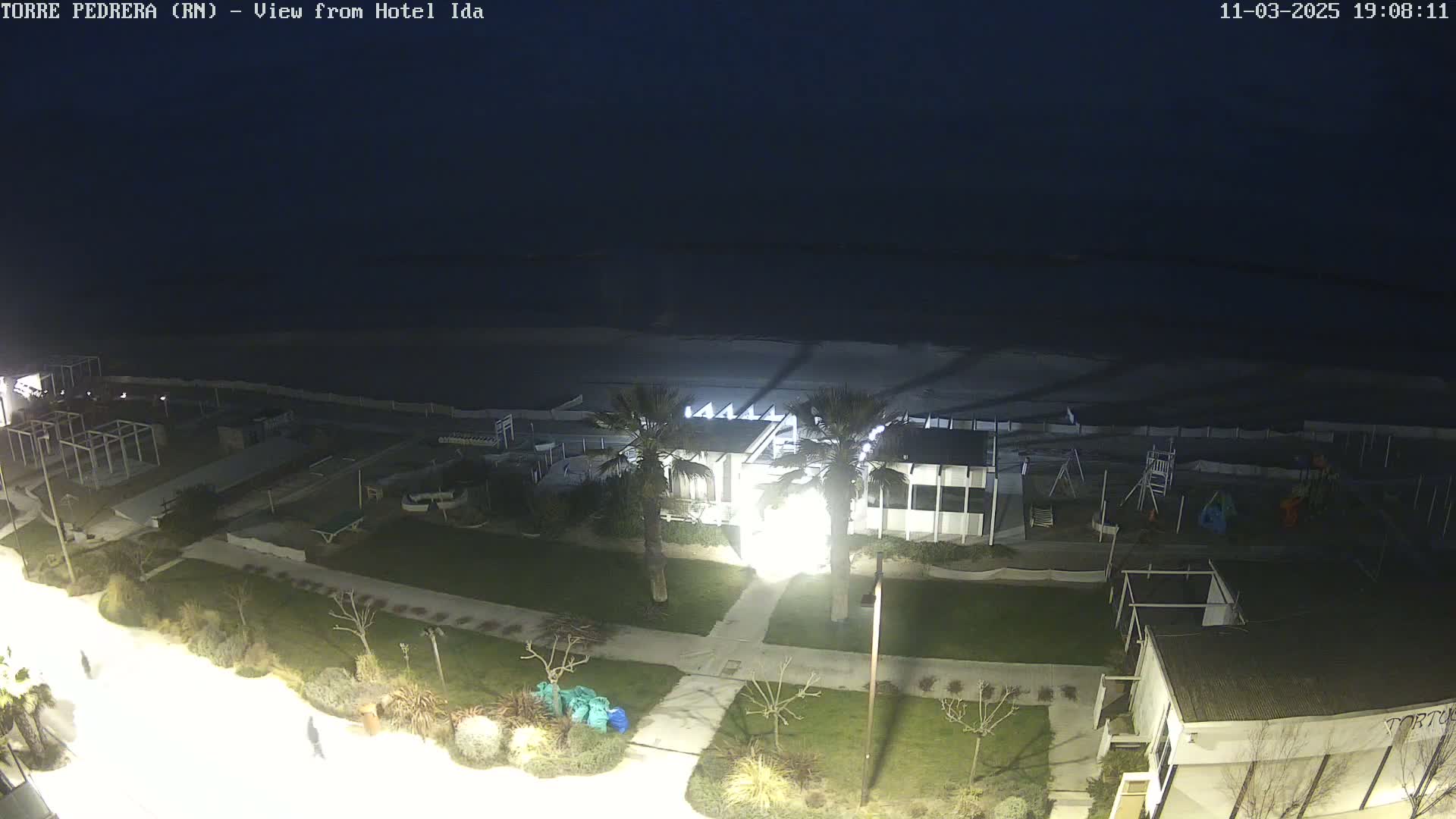 A nighttime view of a beachside area shows a mostly dark beach, a building with lights on, a grassy area with landscaping, and a walkway.