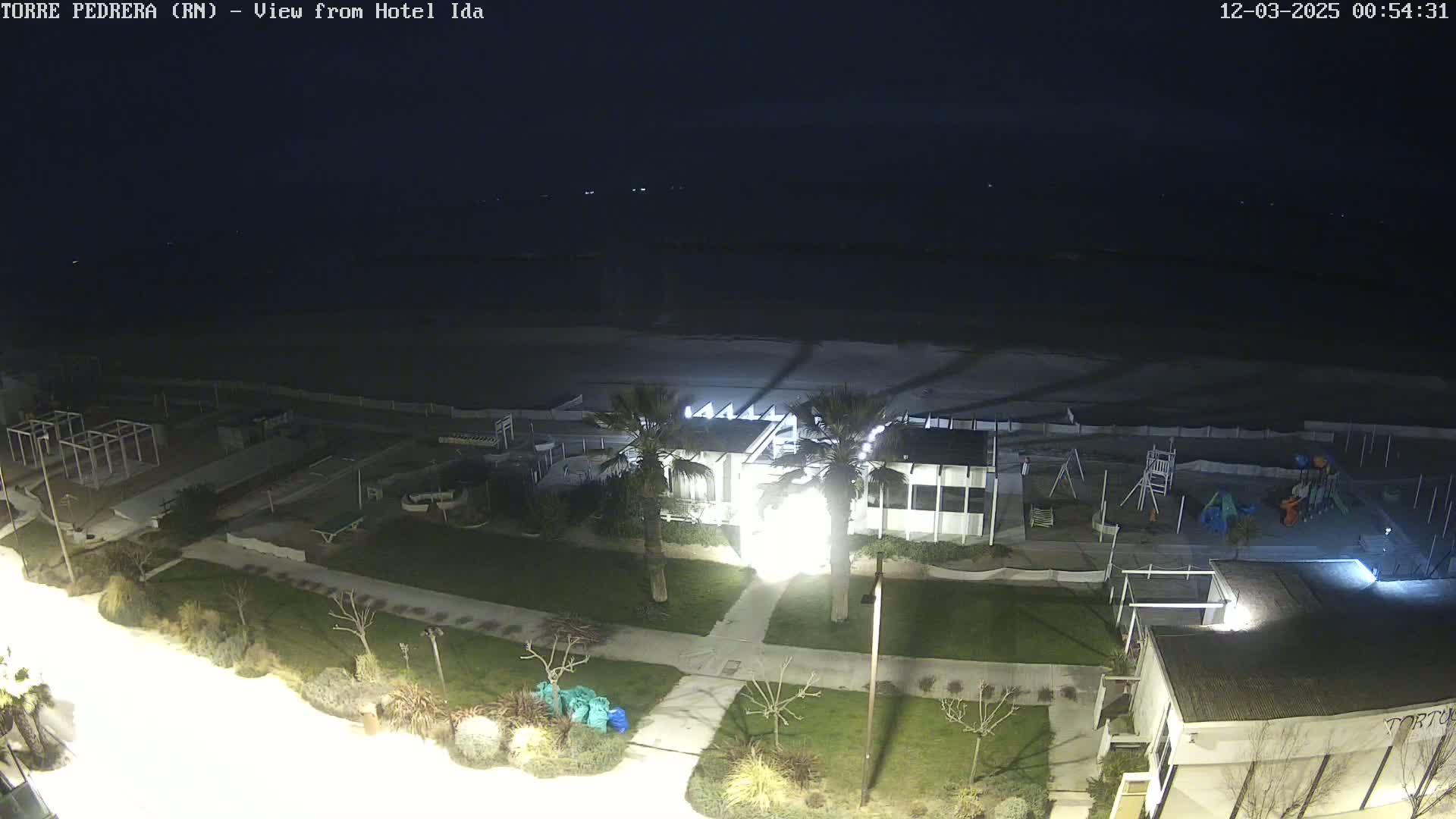 A nighttime view of a beachfront area shows a building, a playground, and a dark sandy beach under a clear, dark sky.