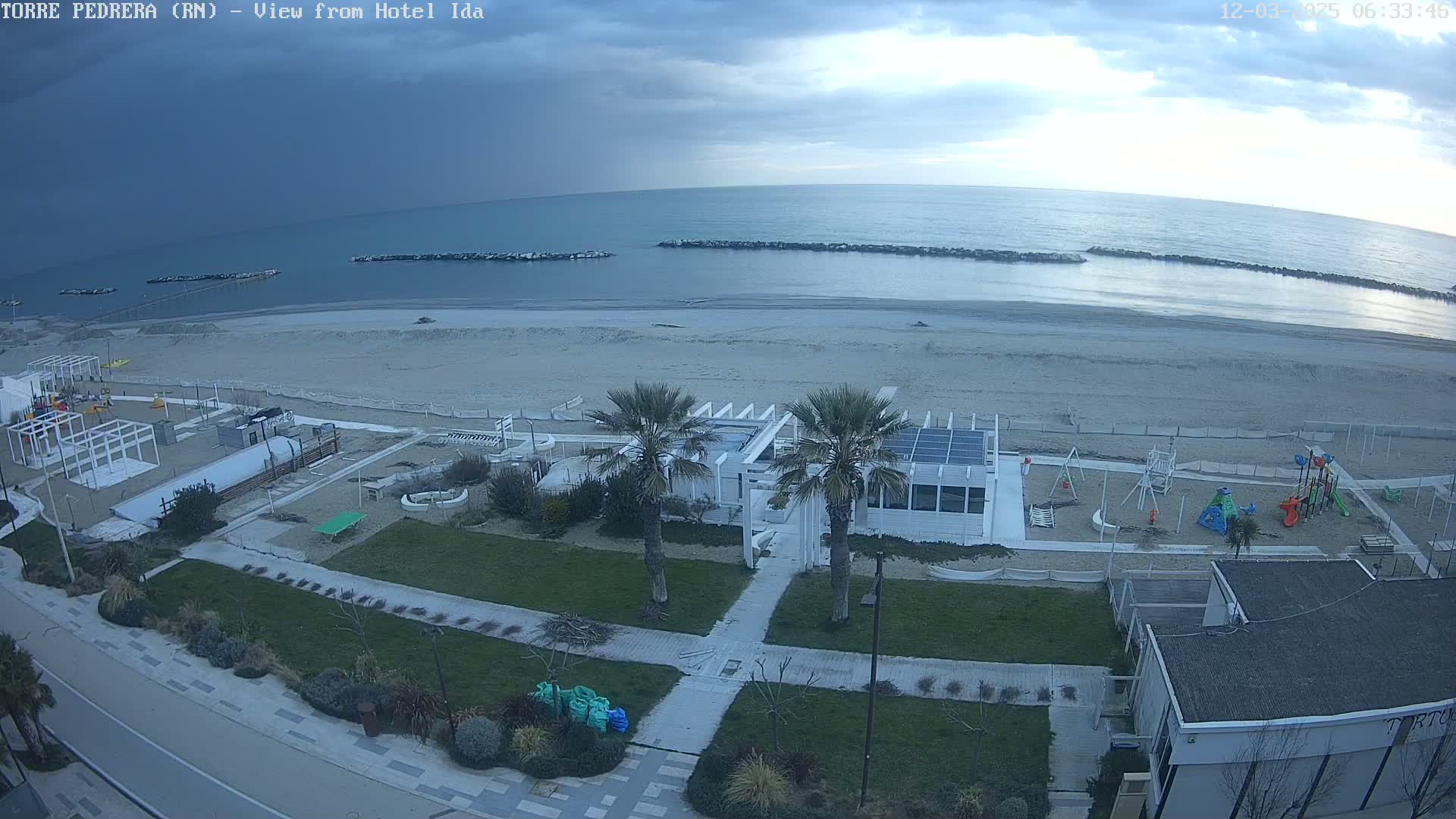 A beach with cloudy skies, a white beachfront building, a playground, and palm trees is viewed from above.