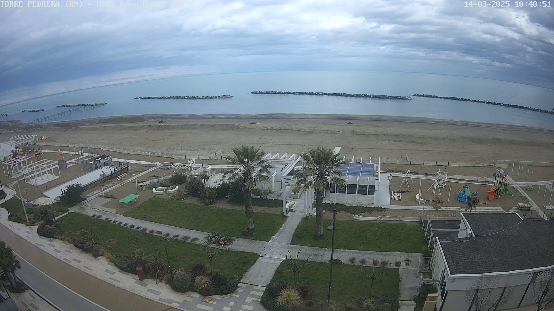 An overhead view of a beach with overcast skies shows a sandy beach, calm sea with breakwaters, a playground, and a white building with palm trees situated near a paved walkway.