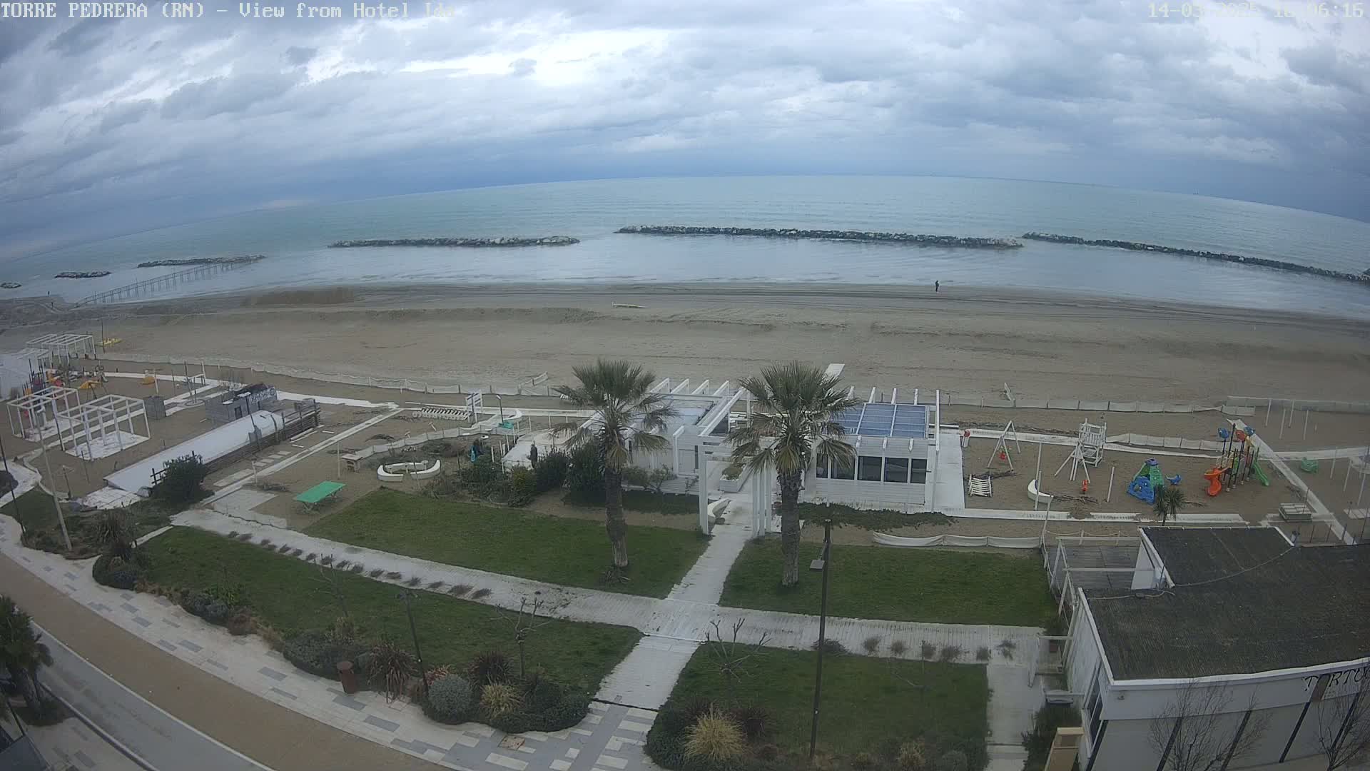 An overcast day reveals a sandy beach with a playground, a beachfront restaurant, and palm trees, all situated near a calm sea protected by breakwaters.