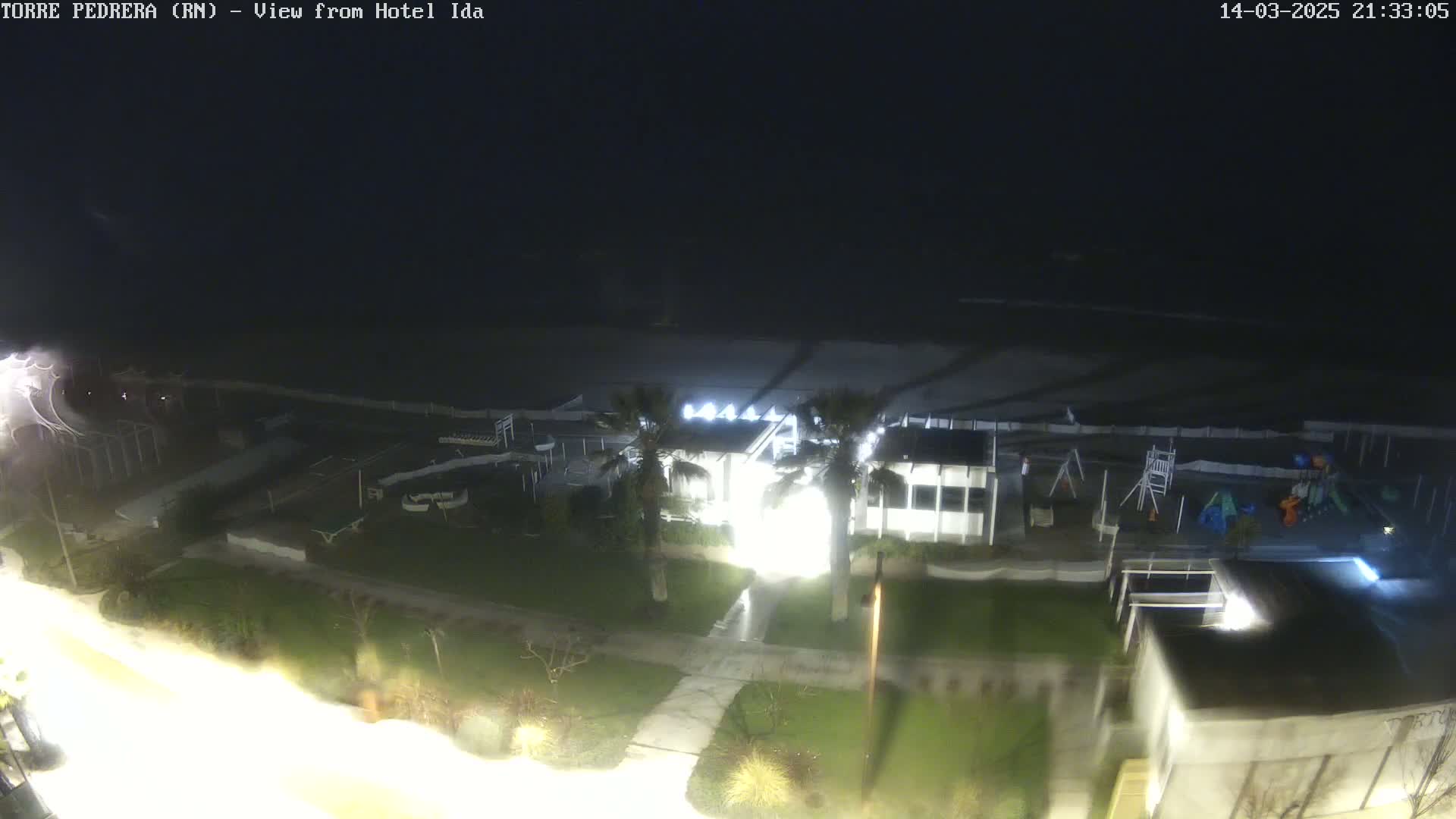 A nighttime, high-angle view shows a beachside building with lights on, a playground, and a dark ocean under a cloudy sky.