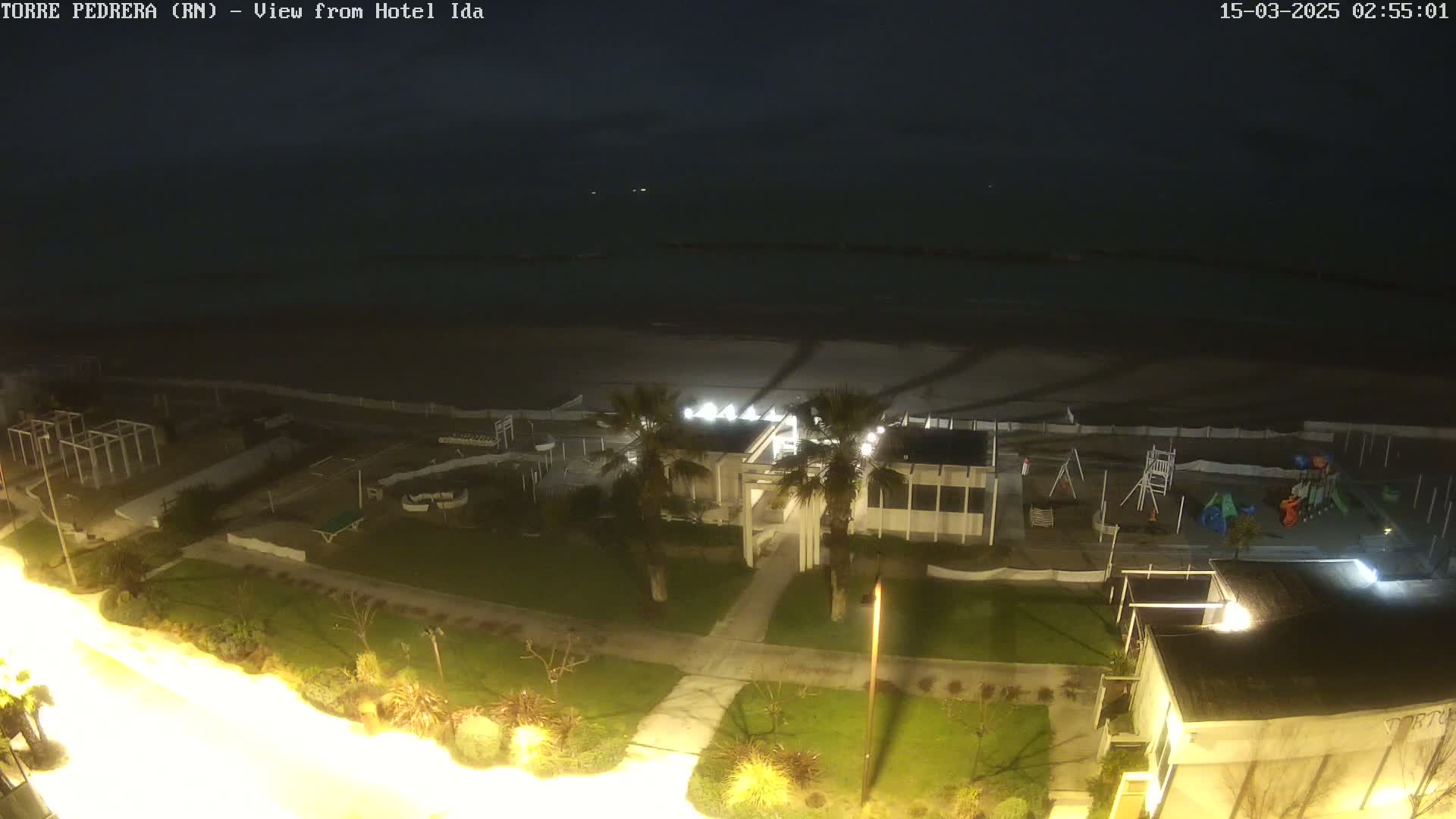 A nighttime, overhead view of a beachside area with a playground, palm trees, and a building, under a dark sky.