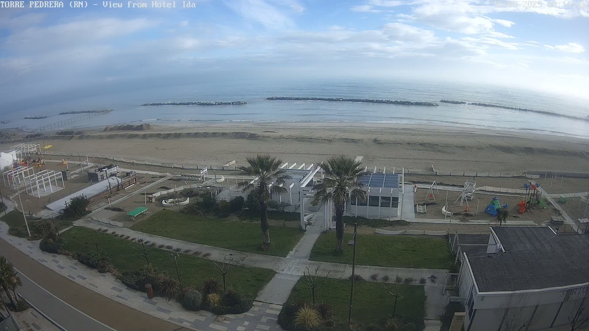An aerial view shows a sandy beach with a calm ocean under a mostly clear sky, a beachfront playground, and a white building near palm trees.