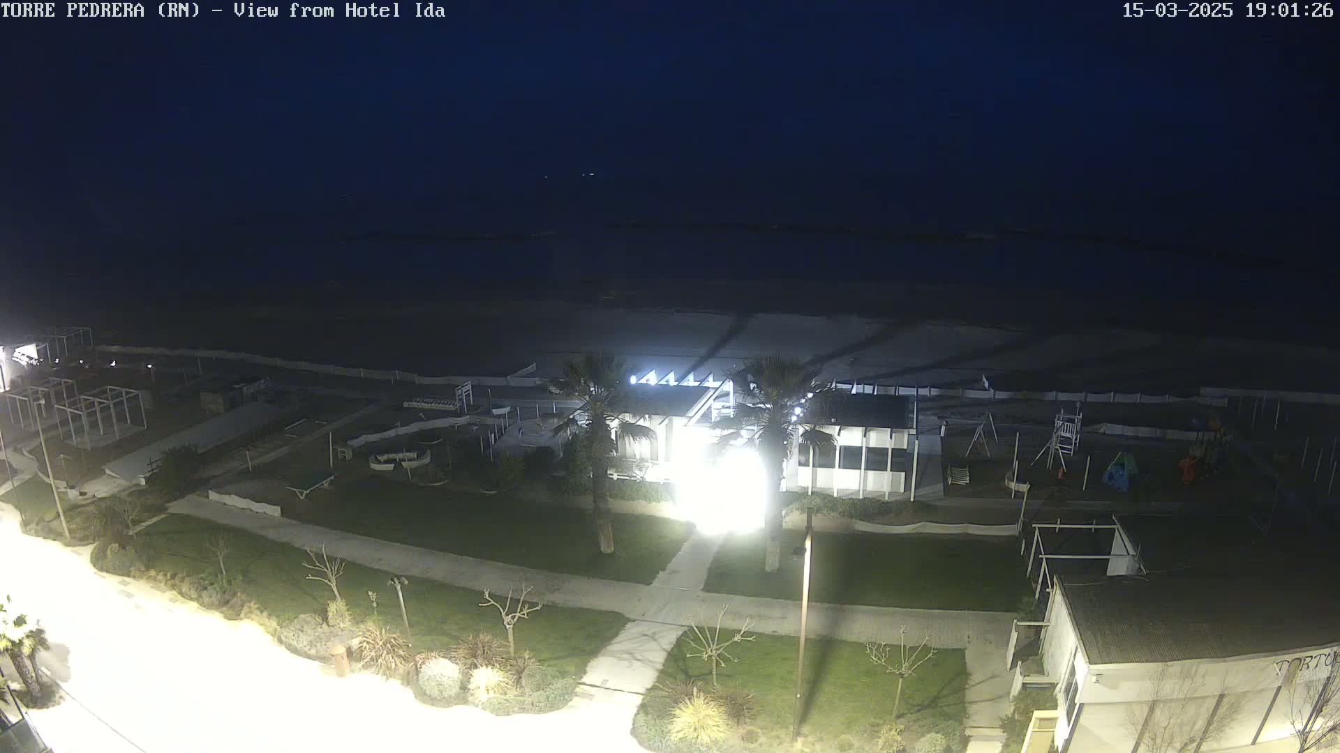 A nighttime, aerial view shows a beach, a beachfront building with lights on, and a grassy area with landscaping, under a dark sky.