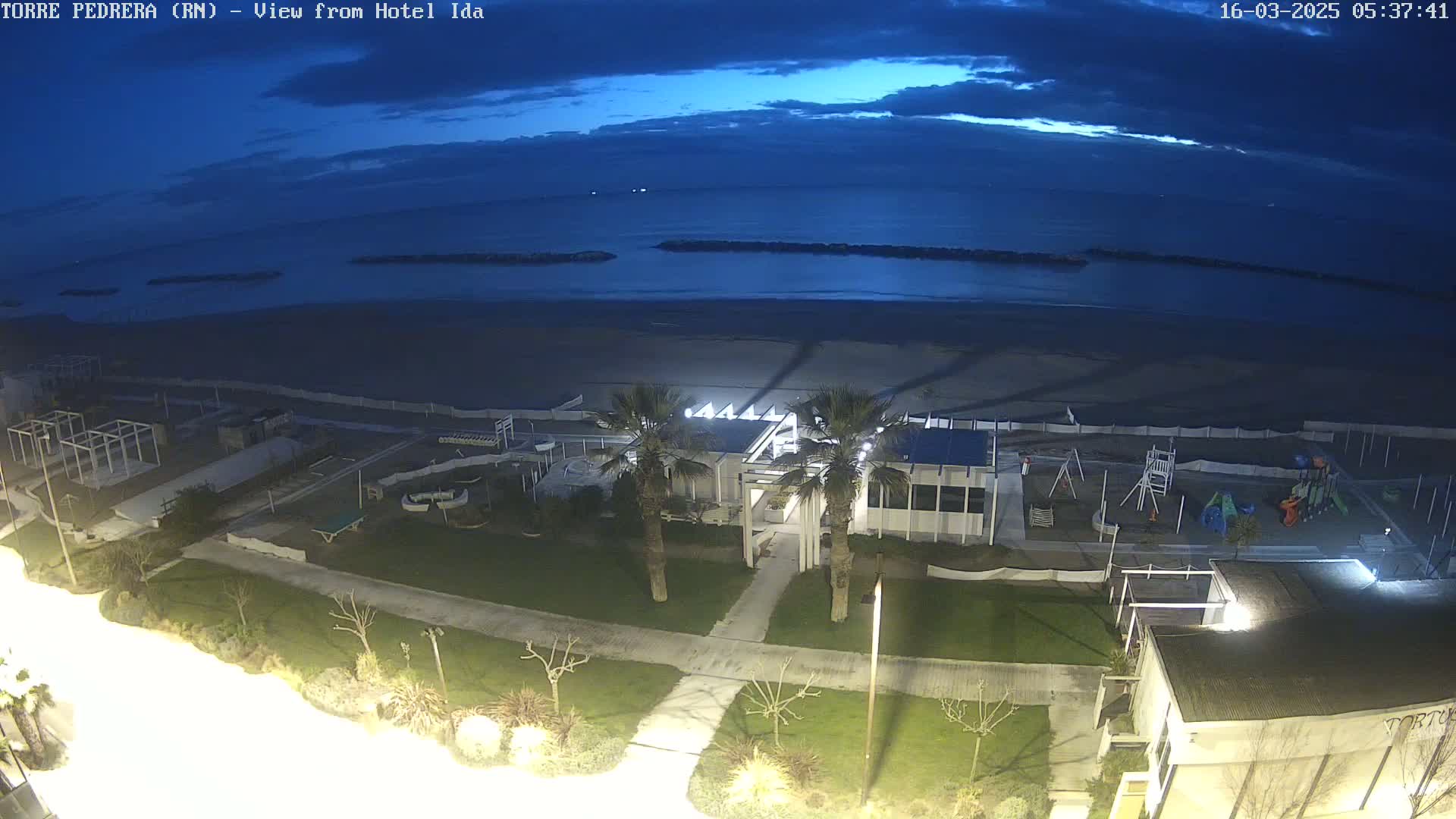 A beach at twilight, with a building, playground, and palm trees visible, under a mostly clear, dark blue sky.