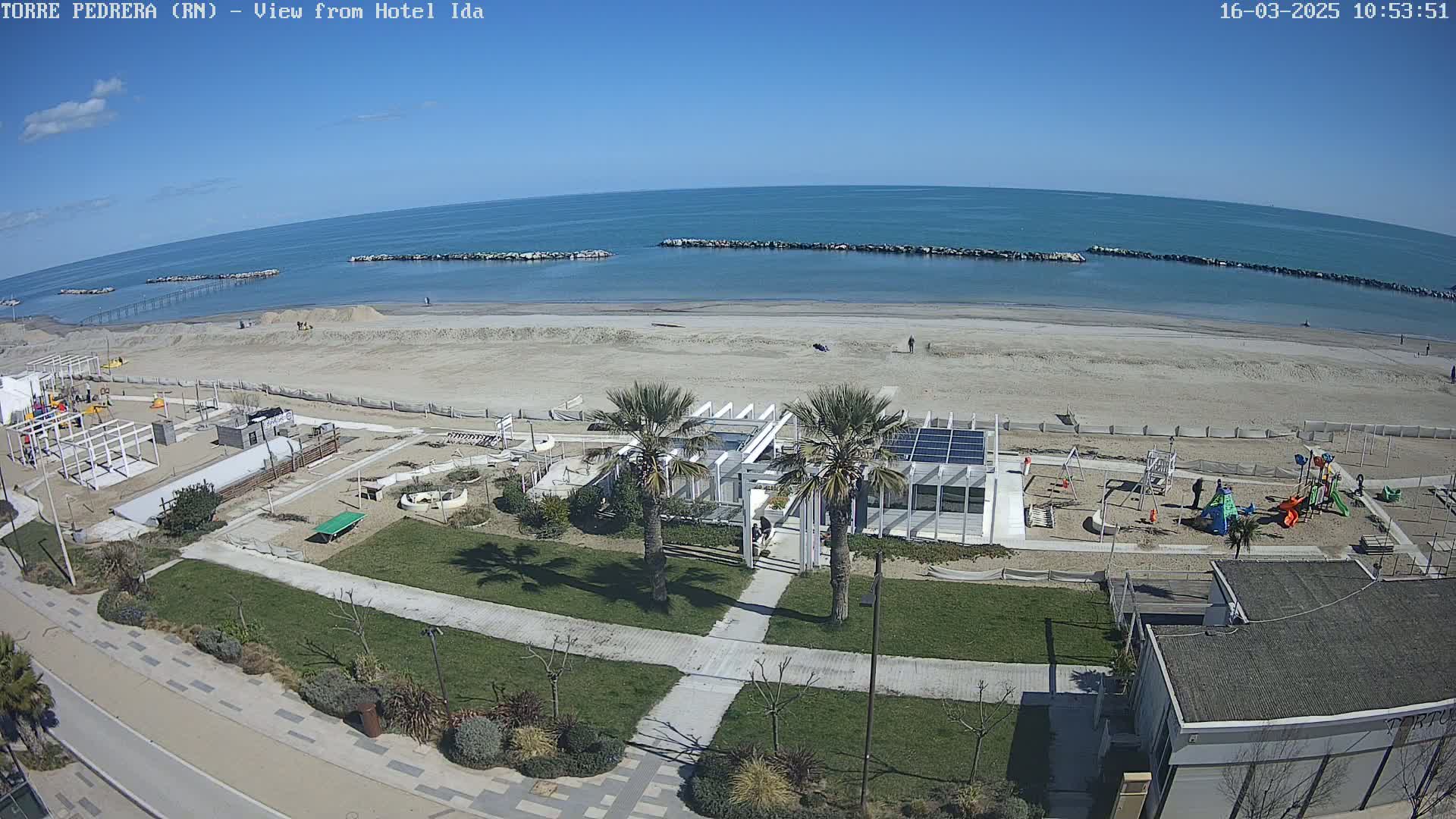 An aerial view shows a sandy beach with calm blue water under a clear blue sky, adjacent to a beachfront area with grassy lawns, a playground, and a white building with palm trees.