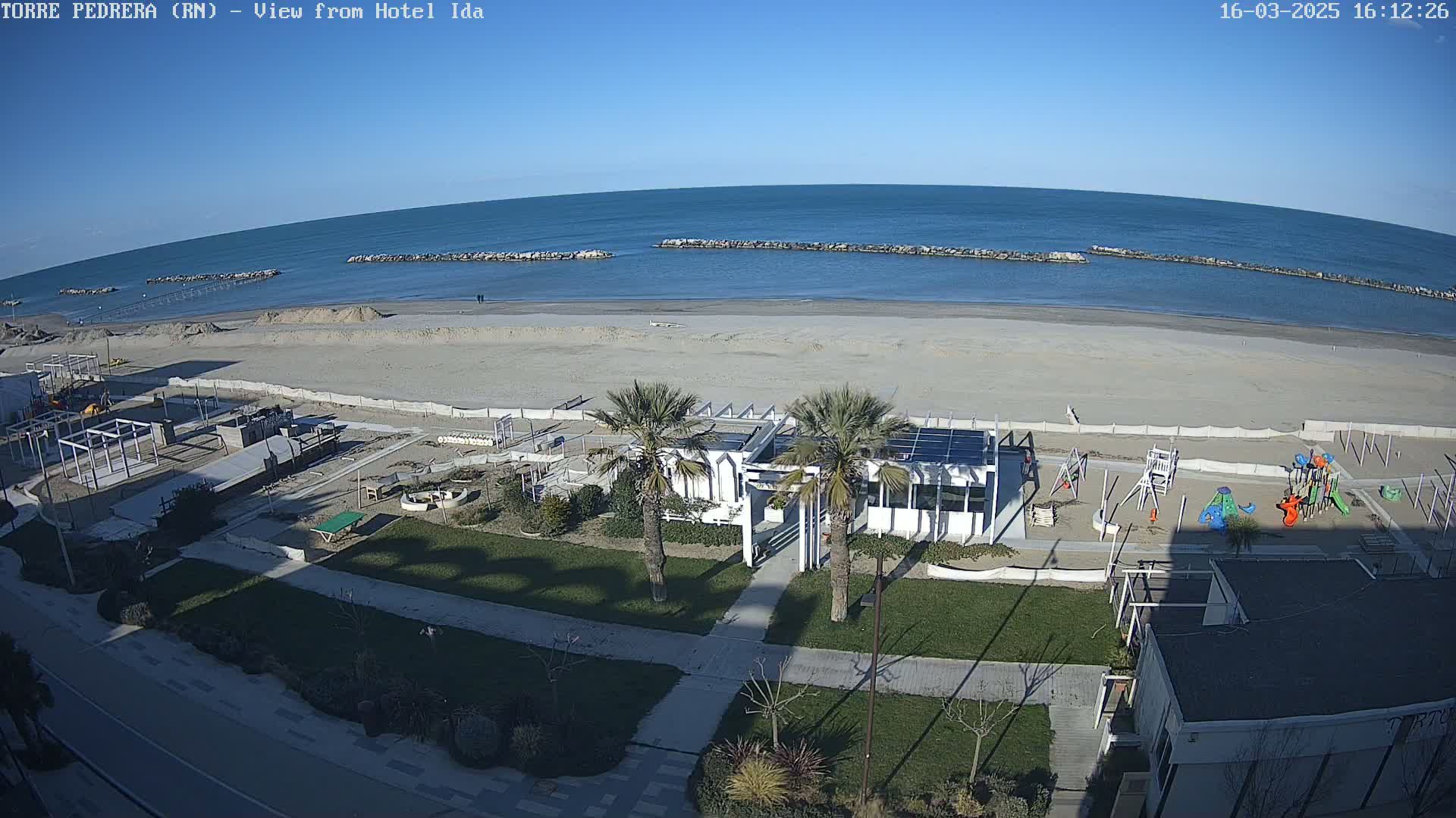A sandy beach with calm ocean water under a clear blue sky, featuring a beachfront building with palm trees and a children's playground nearby.