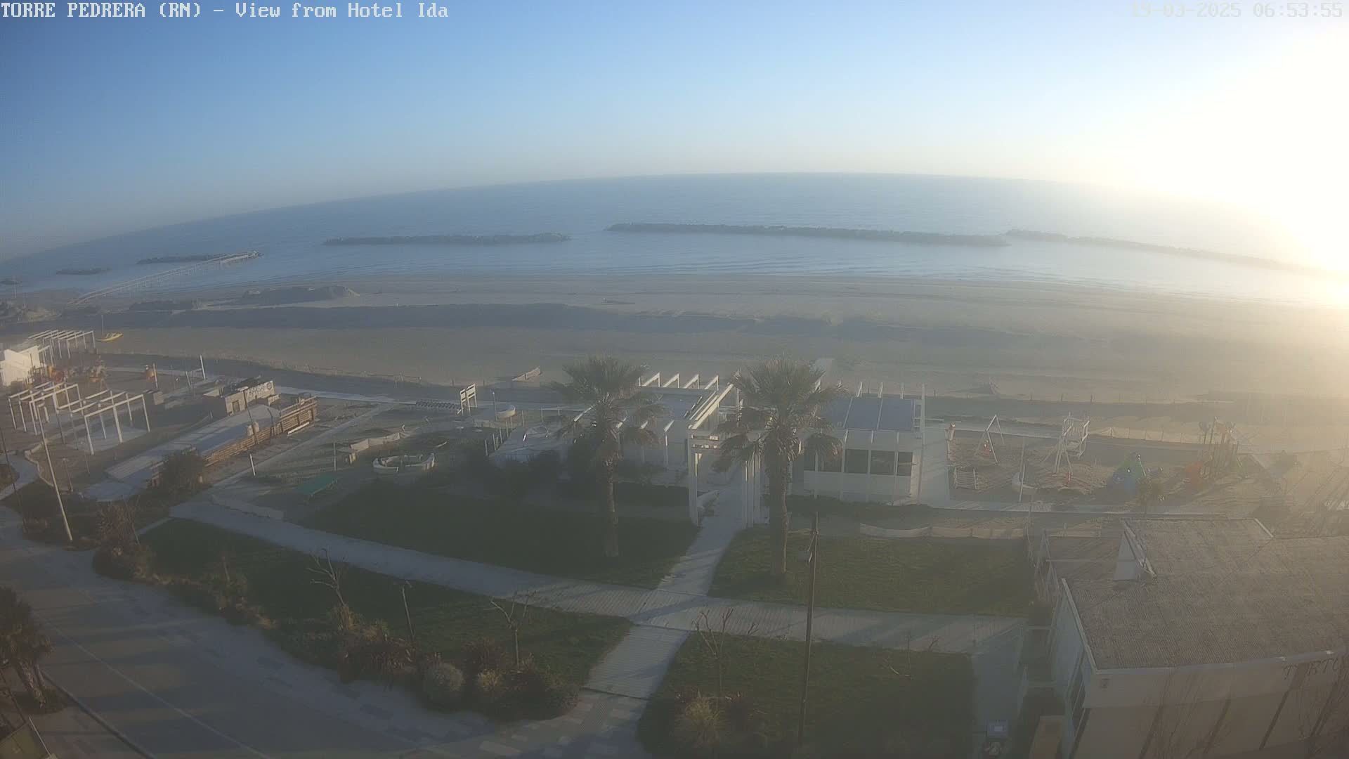 An aerial view of a beach with calm water under a hazy sky shows a sandy beach, a playground, and a white building near palm trees.