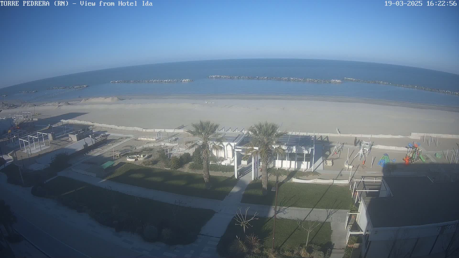 A beach with calm, clear water under a sunny sky is seen from an elevated vantage point, showcasing a sandy shore with a playground and a beachfront building.