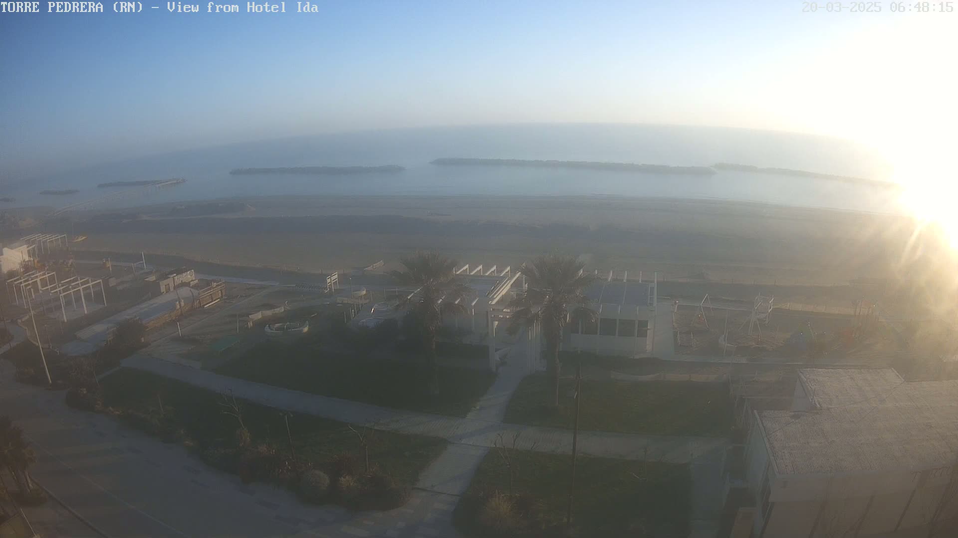 A hazy sunrise illuminates a beach scene with a building, palm trees, and a playground near a calm ocean with breakwaters visible in the distance.