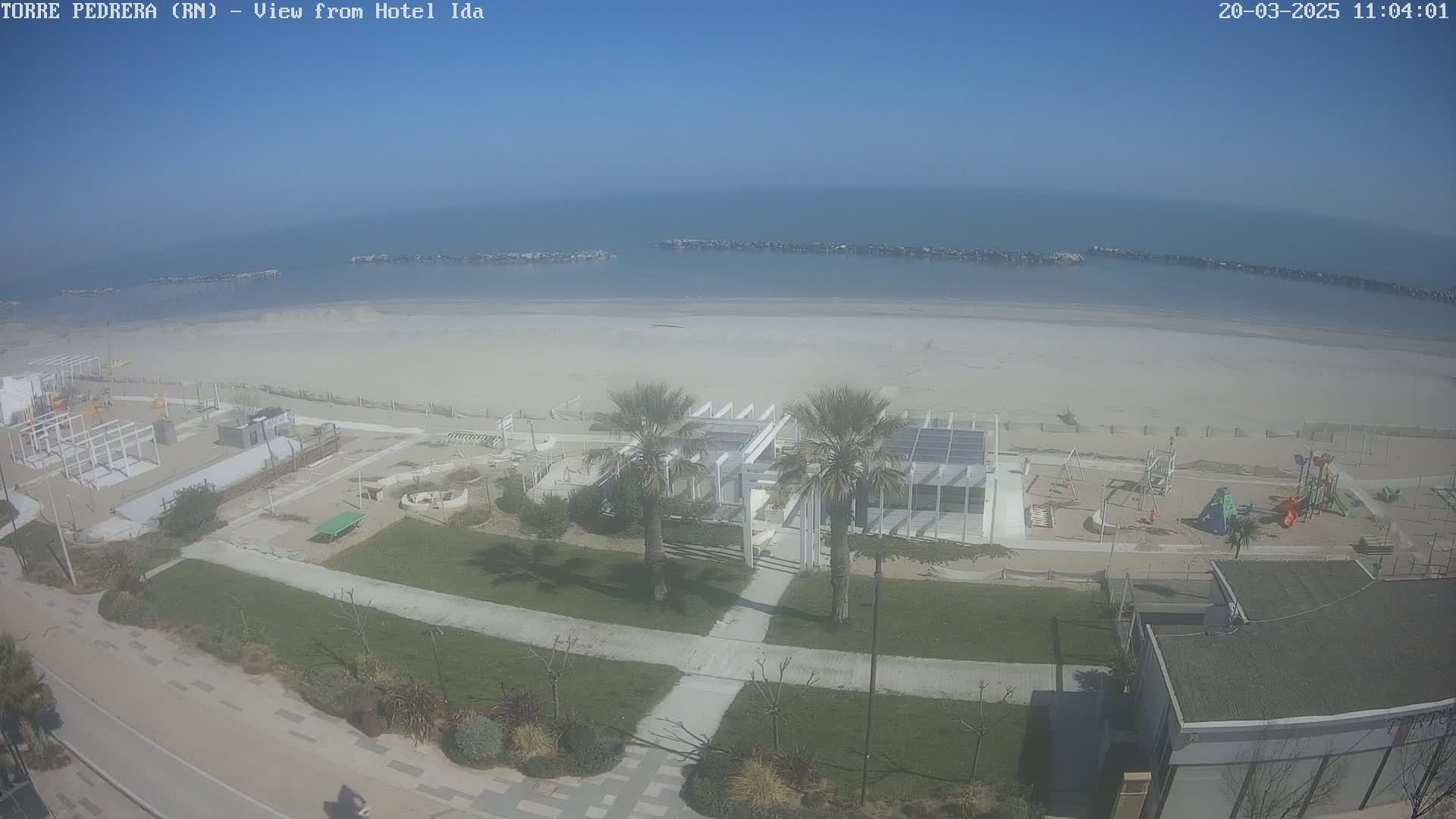 An aerial view shows a sandy beach with calm ocean waters under a clear blue sky, adjacent to a grassy area with a playground and a modern white structure.