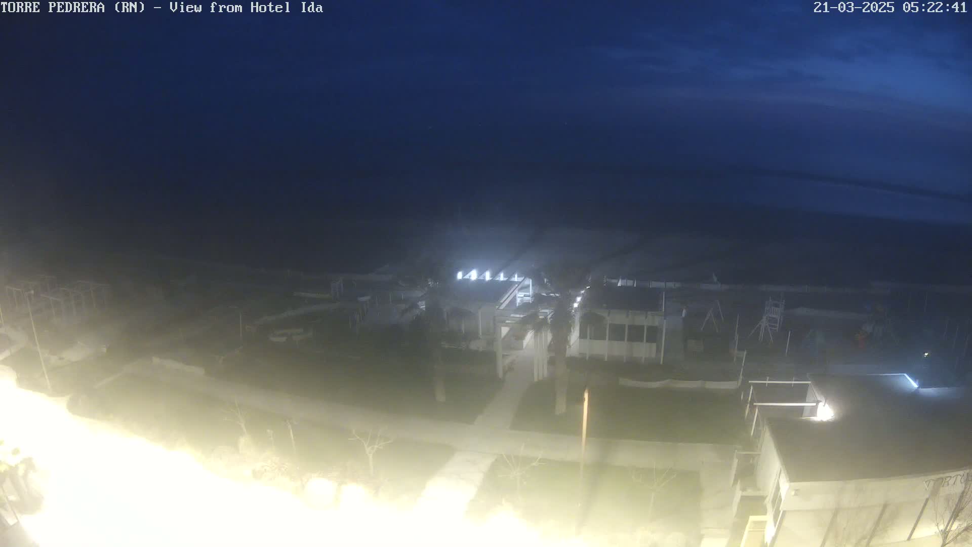 A nighttime, dimly lit view of a coastal area shows buildings and a playground near a dark body of water under a mostly clear, dark blue sky.