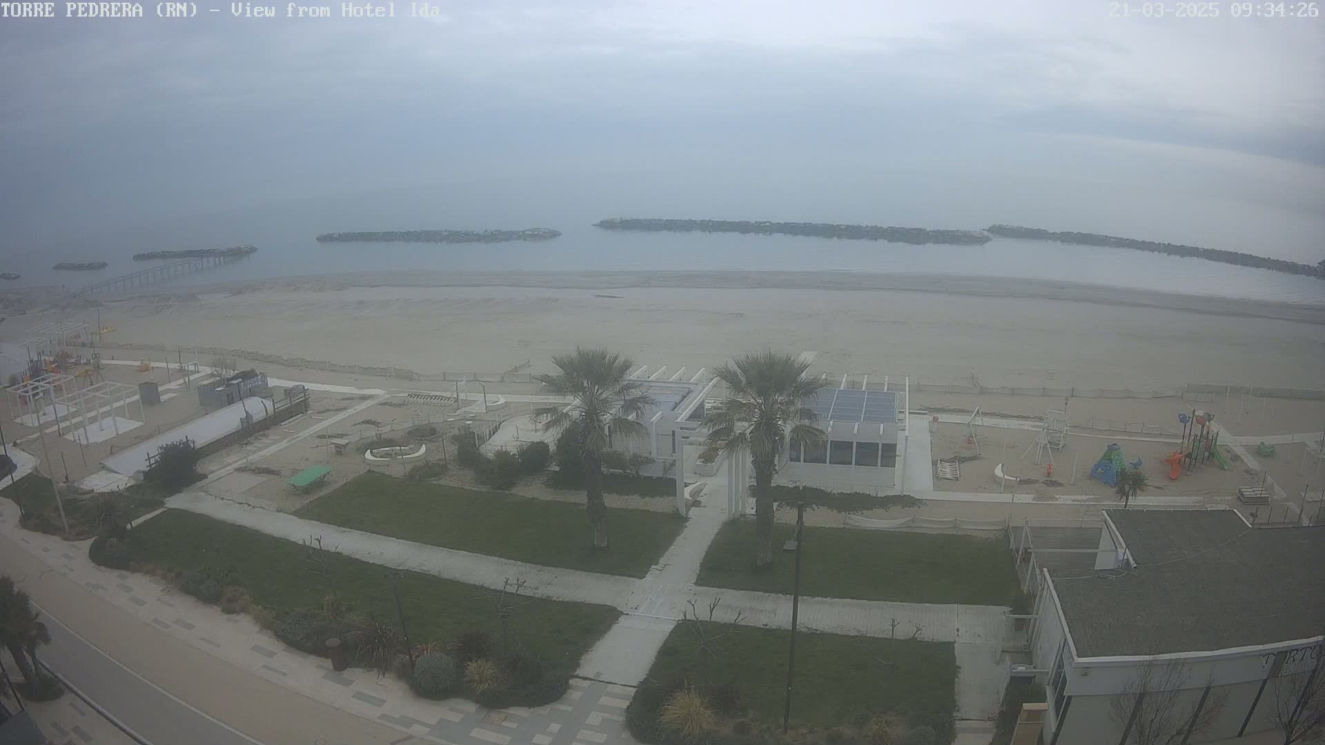 An overcast day reveals a beach with a playground, some buildings, and palm trees, all seen from an elevated vantage point.