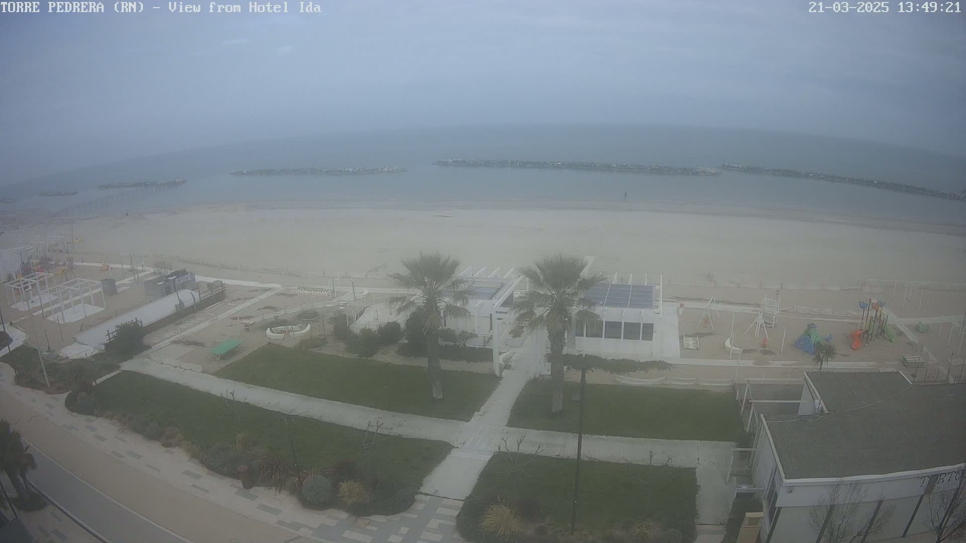 A hazy, overcast day reveals a beach scene with a sandy shore, calm water, a beachfront building with palm trees, playground equipment, and paved walkways.