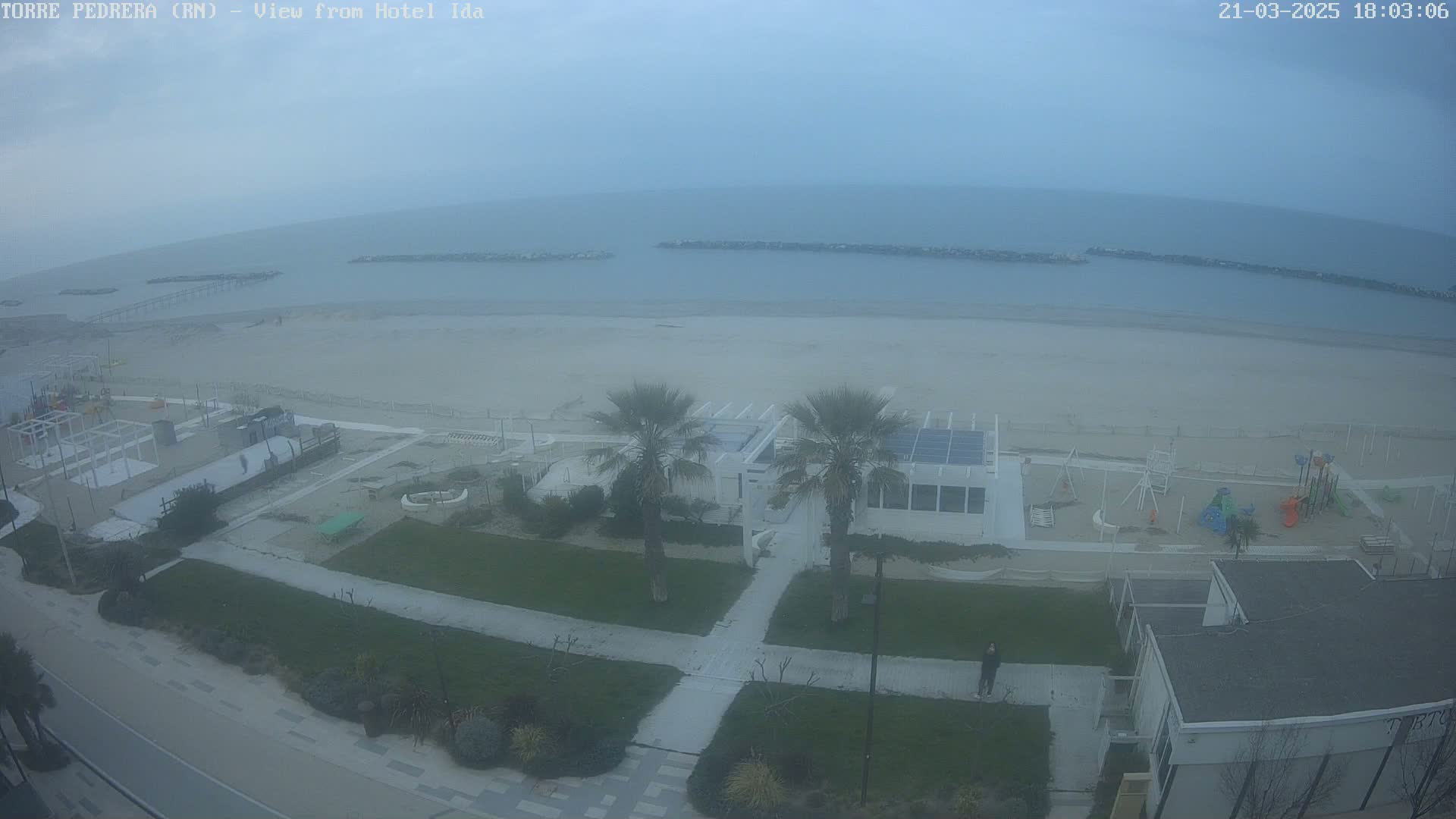 A hazy, overcast day shows a beach with a playground, palm trees, and buildings near the water.