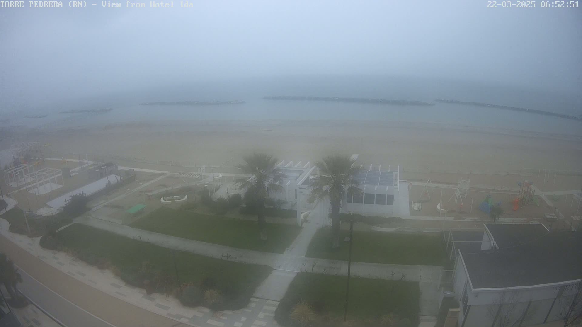 A foggy beach scene shows a sandy beach, a walkway, some buildings, palm trees, and playground equipment.