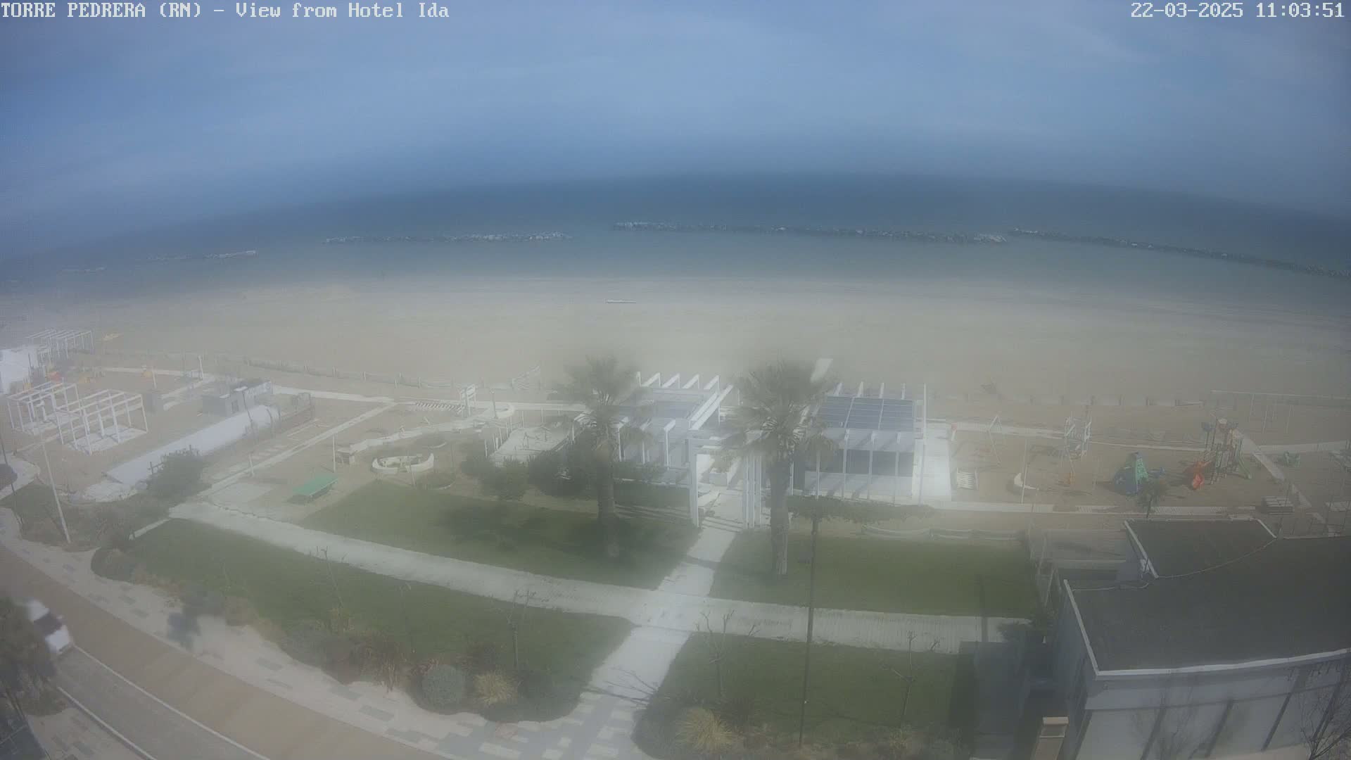 A hazy beach scene shows a sandy shore, calm ocean, beachfront buildings, and palm trees under a mostly clear sky.