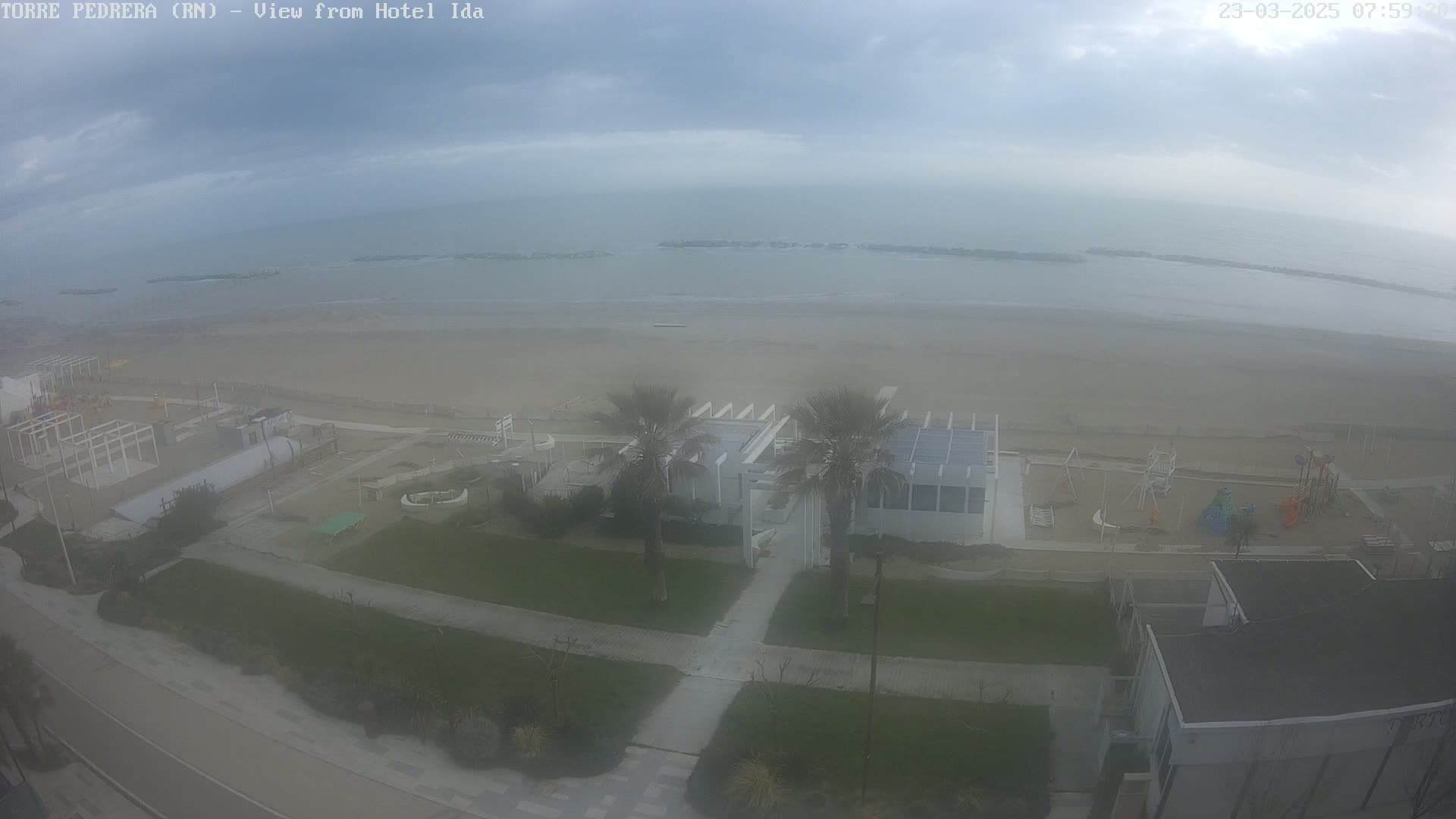 An overcast day reveals a beach with a playground, palm trees, and a white building near the water.