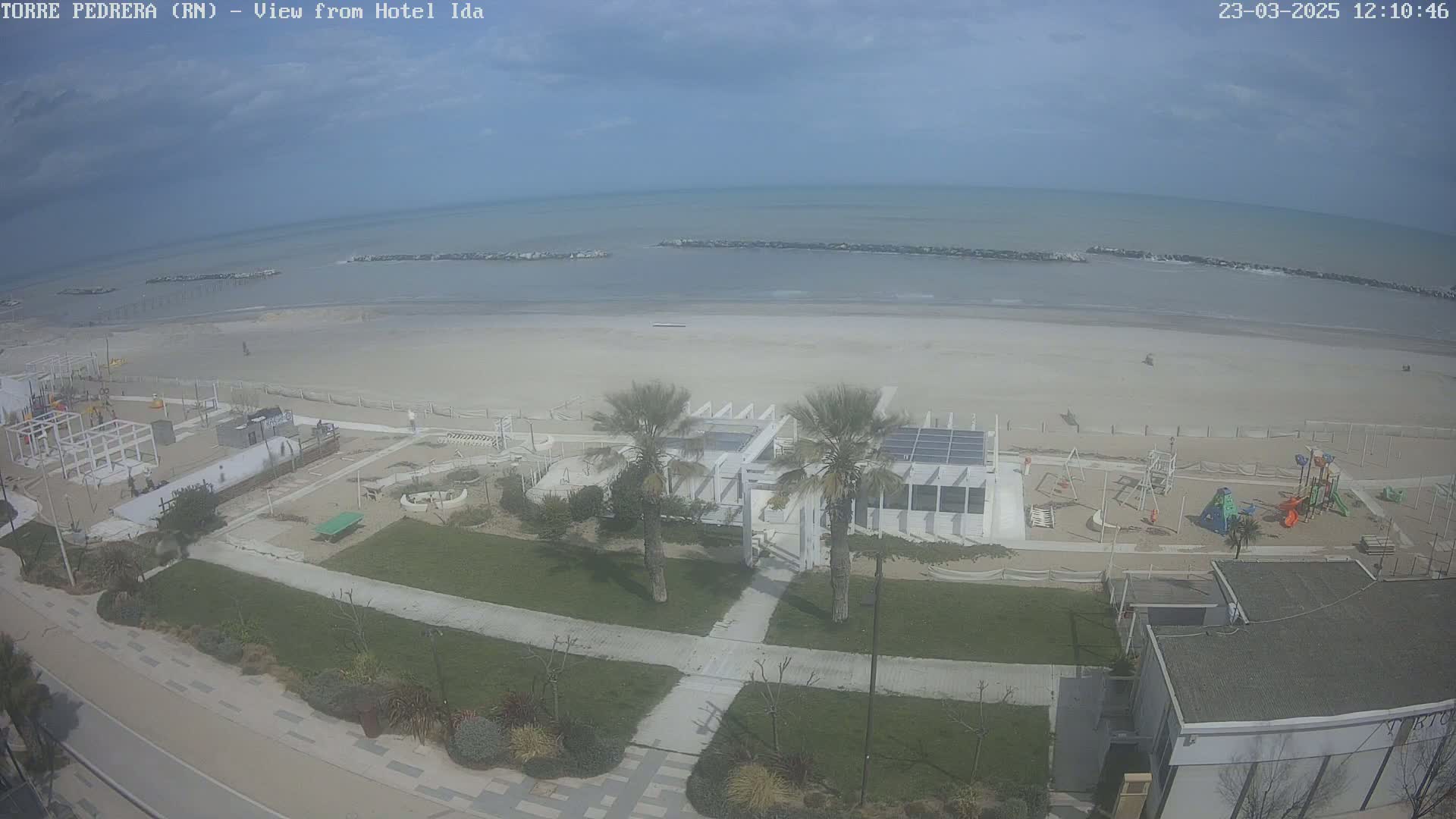 An aerial view shows a beach with calm water, a beachfront building with palm trees, and a playground under a mostly clear sky.