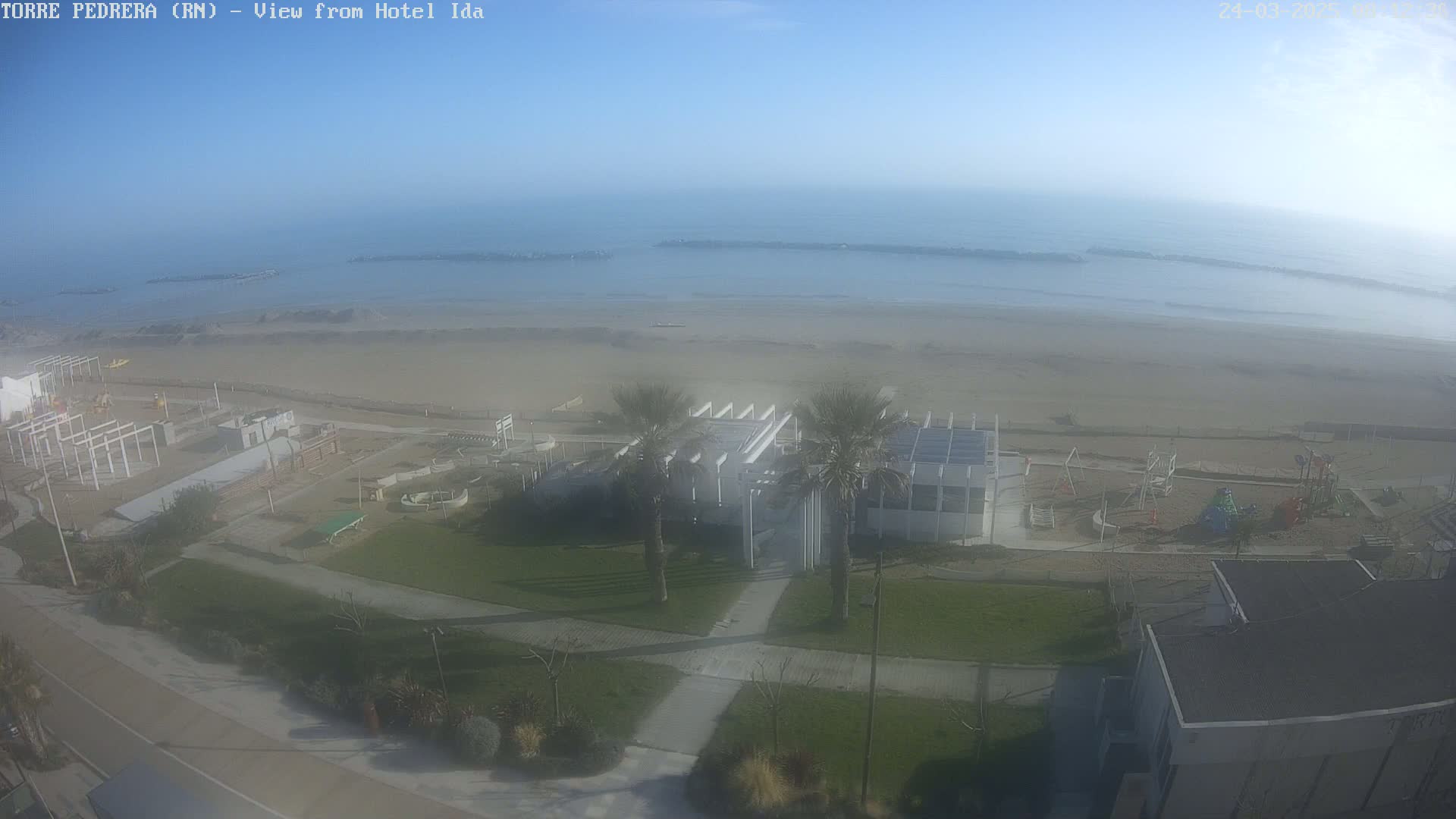 A hazy, sunny day overlooks a beach with a sandy shoreline, a playground, a white building with palm trees, and a roadway.