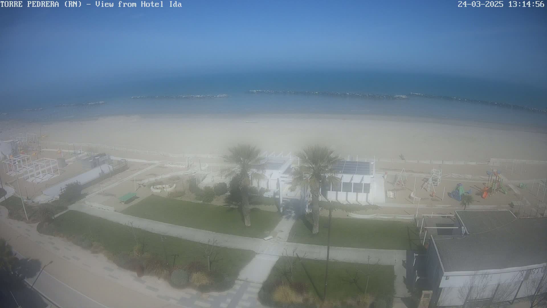 An aerial view shows a beach with calm water under a clear, bright sky, featuring a beachfront playground, palm trees, and a building near the shore.
