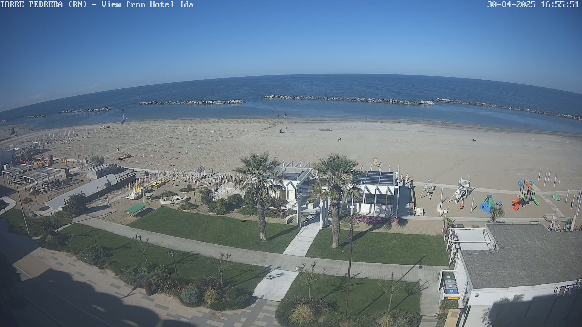 A beach with calm, clear water under a sunny sky, featuring a beachfront building, playground, and palm trees.