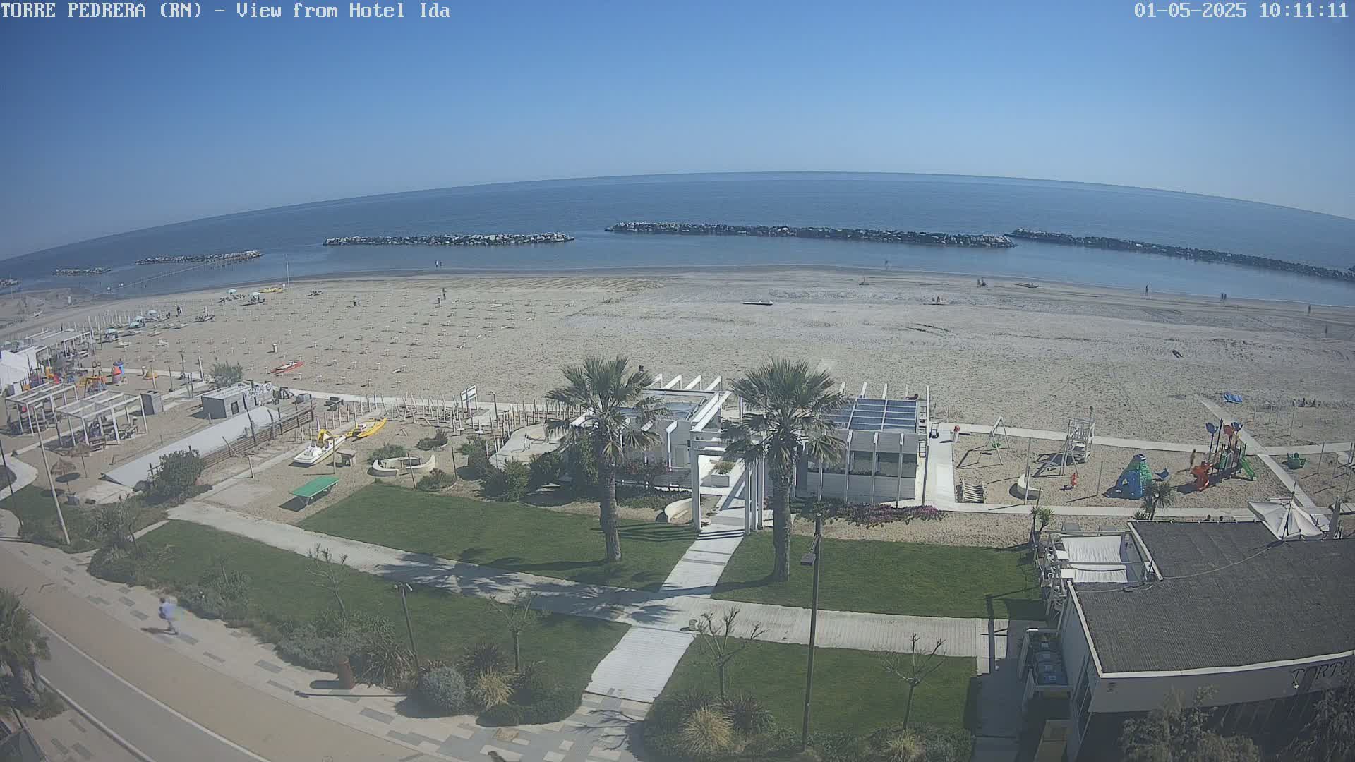 A beach with a playground and beachfront buildings is seen from above on a sunny day.