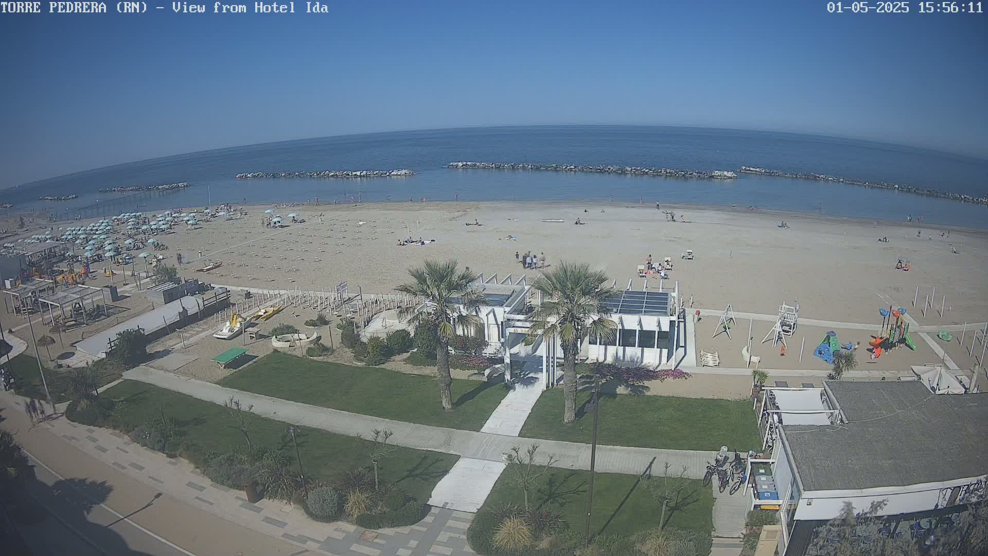 A sandy beach with scattered beachgoers, lined with umbrellas and beachside amenities under a clear blue sky.