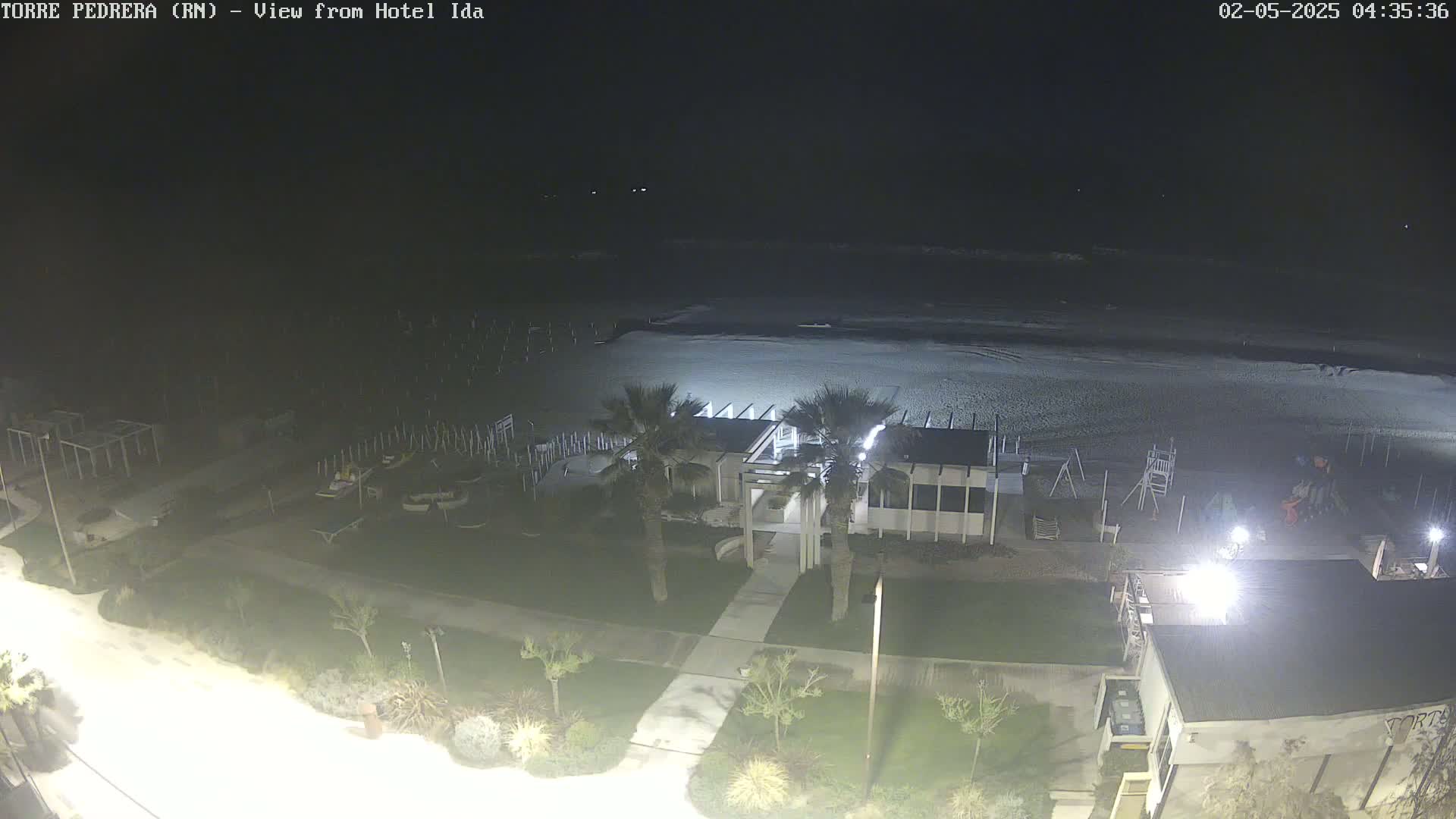 A nighttime, high-angle view shows a mostly empty beach, a beachfront building with lights, and surrounding landscaping.