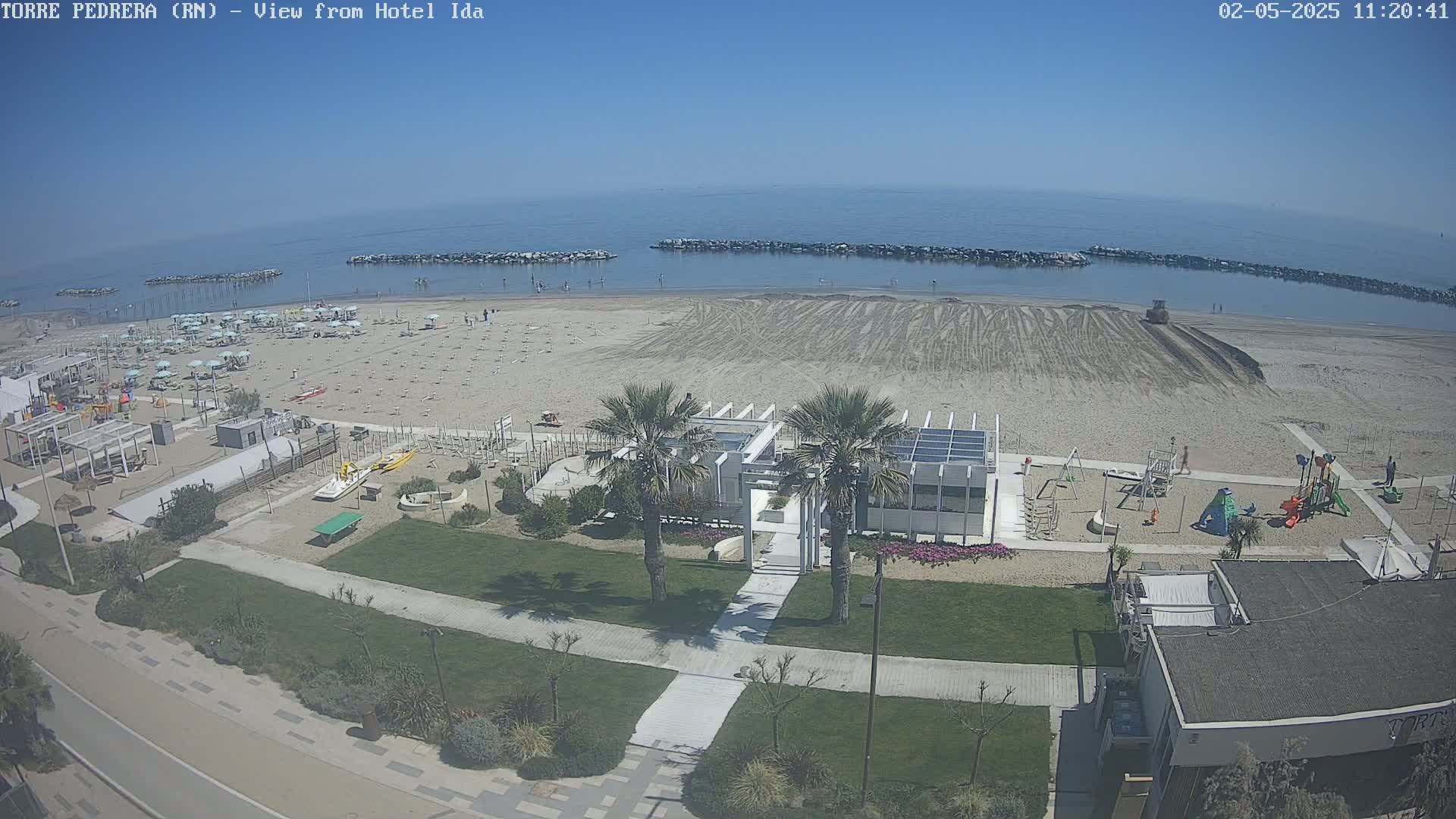 A beach scene on a sunny day shows a sandy beach with beach umbrellas, a playground, and palm trees near a building, all next to calm blue water.