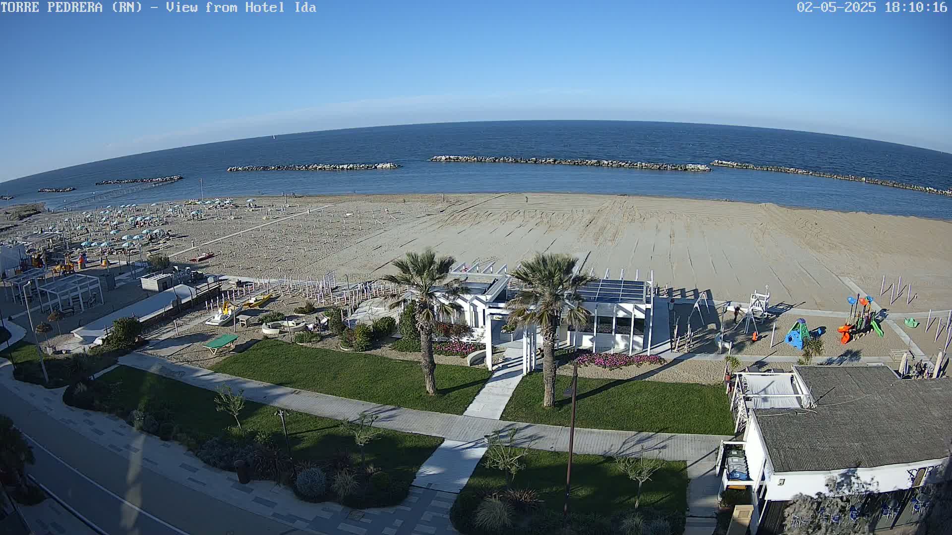 A beach with a calm sea under a clear blue sky shows a sandy beach with a playground, palm trees, and beach umbrellas, near buildings and a walkway.