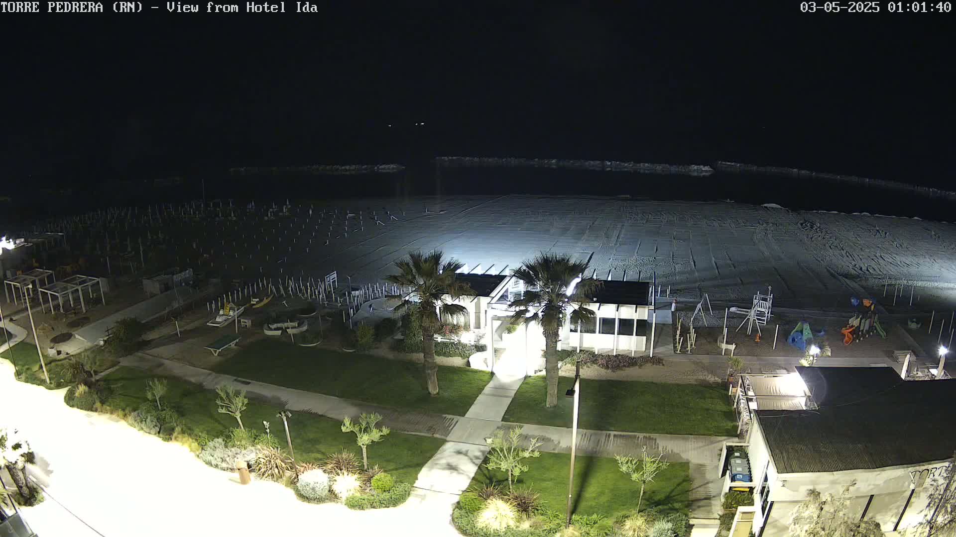 A nighttime, aerial view of a beach with closed beach umbrellas, a playground, and a beachfront building illuminated by lights.
