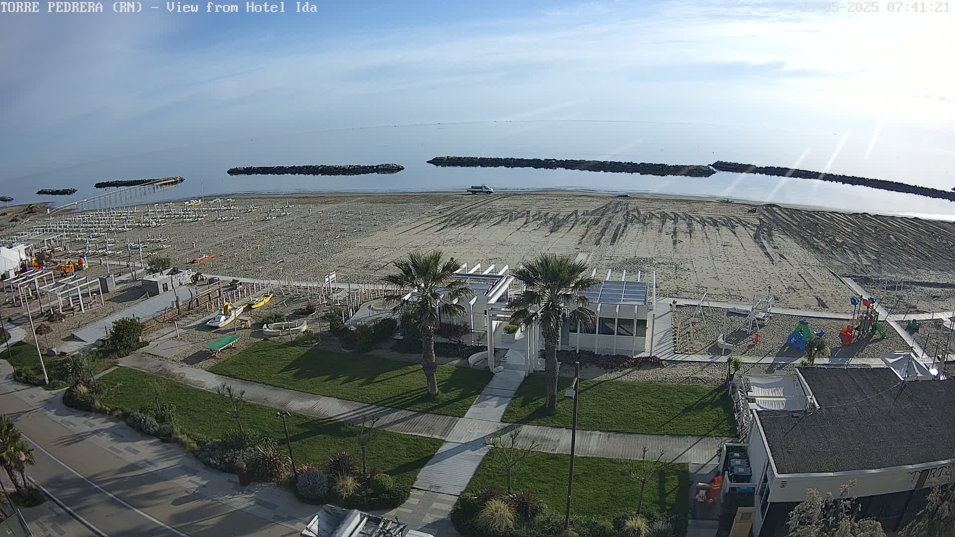 An aerial view of a beach shows rows of beach umbrellas and chairs, a playground, a white building with palm trees, and calm, clear water under a mostly clear sky.