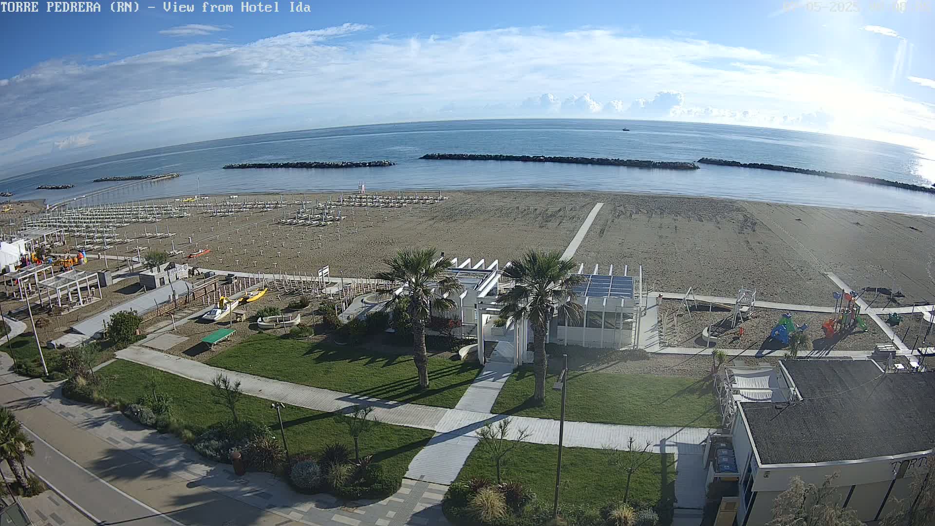 An aerial view shows a sandy beach with rows of beach umbrellas, a playground, and a white building near palm trees, all under a mostly sunny sky.