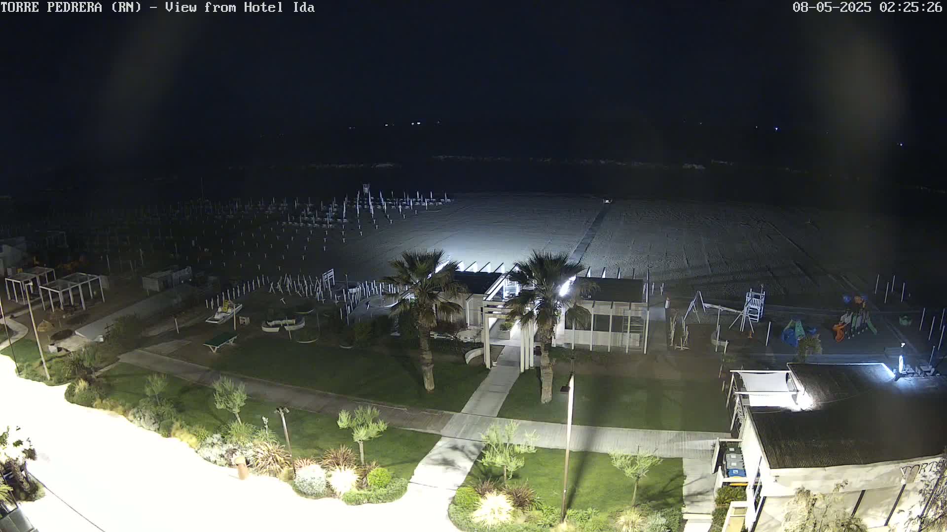 A nighttime aerial view shows a sandy beach with a boardwalk, beach umbrellas, a playground, and a building with palm trees, all illuminated under a dark sky.
