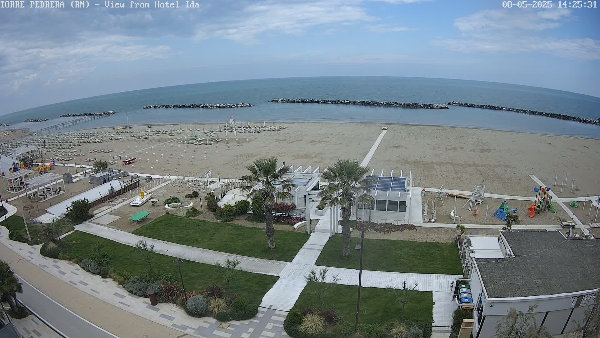 An aerial view shows a beach with rows of beach umbrellas, a playground, and palm trees near a white building, all under a partly cloudy sky.