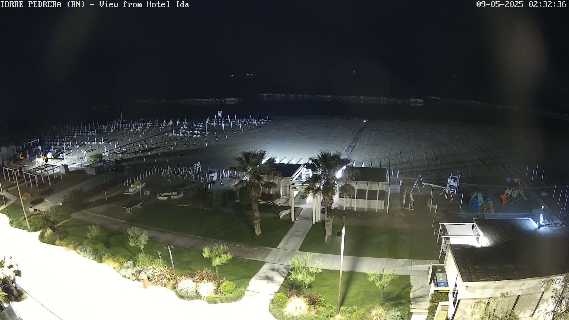 A nighttime view of a sandy beach with beach umbrellas, a playground, and palm trees, overlooking a calm sea under a dark sky.