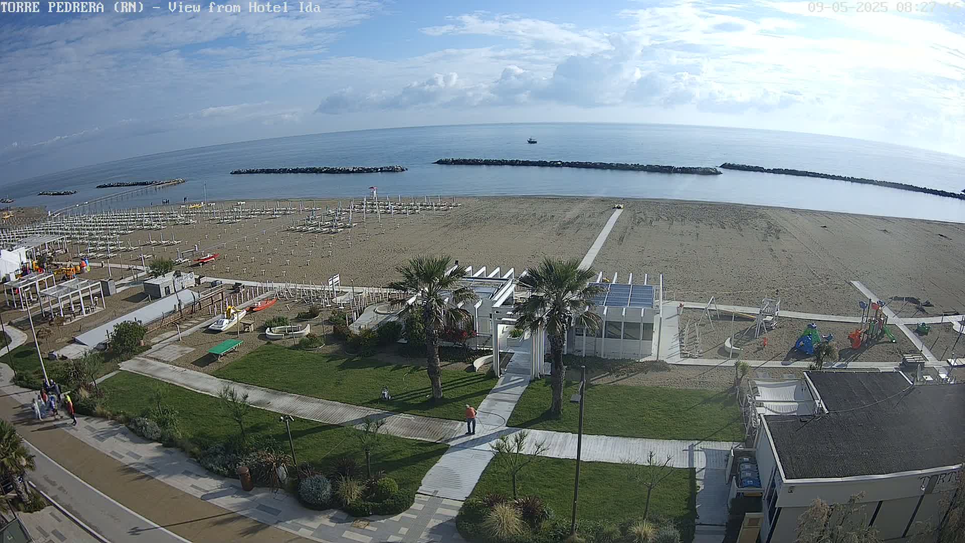 An aerial view shows a sandy beach with rows of beach umbrellas, a playground, and a walkway leading to a white building, all under a partly cloudy sky.