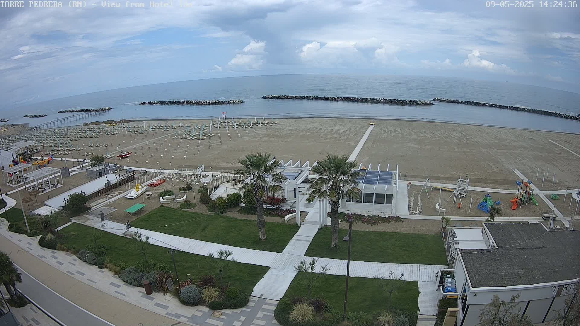 A mostly sunny day at a beach shows rows of beach umbrellas on the sand, a playground, and beachfront buildings with palm trees.