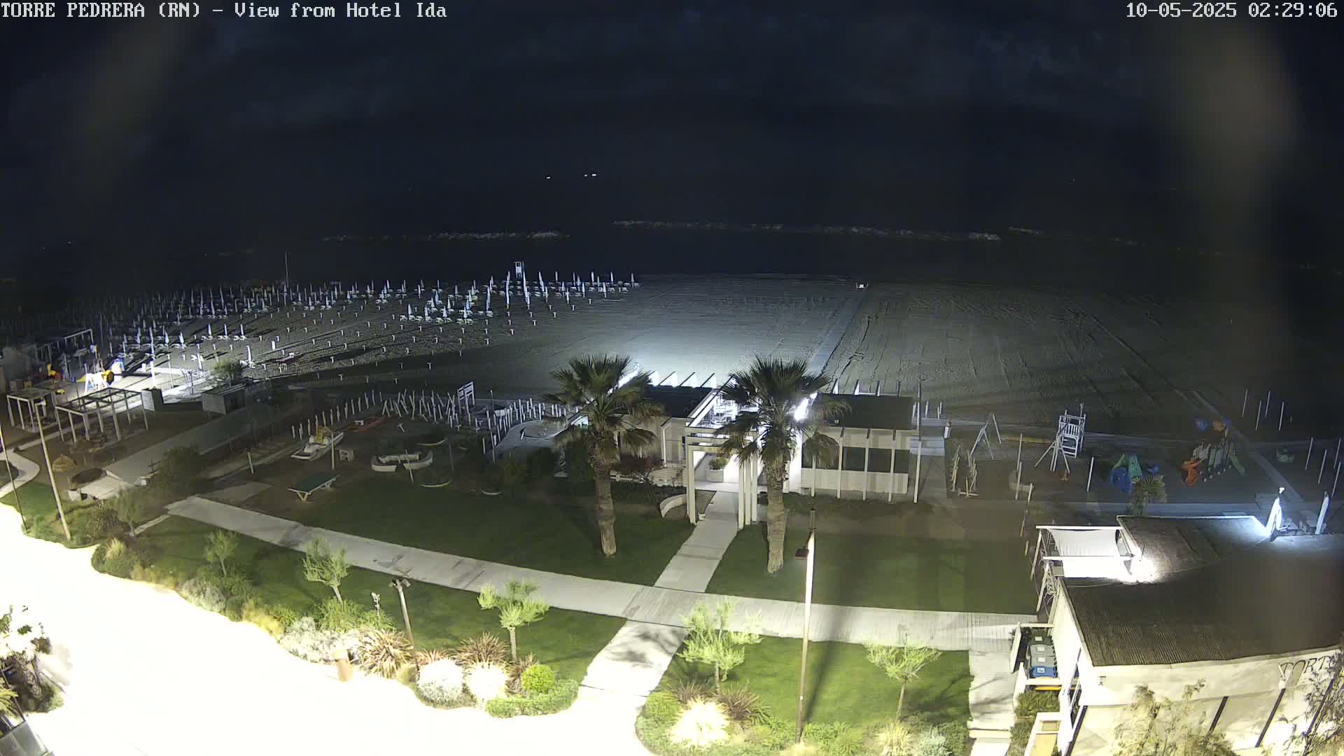 A nighttime aerial view shows a mostly empty beach with rows of closed beach umbrellas, a beachfront building, a playground, and landscaping.